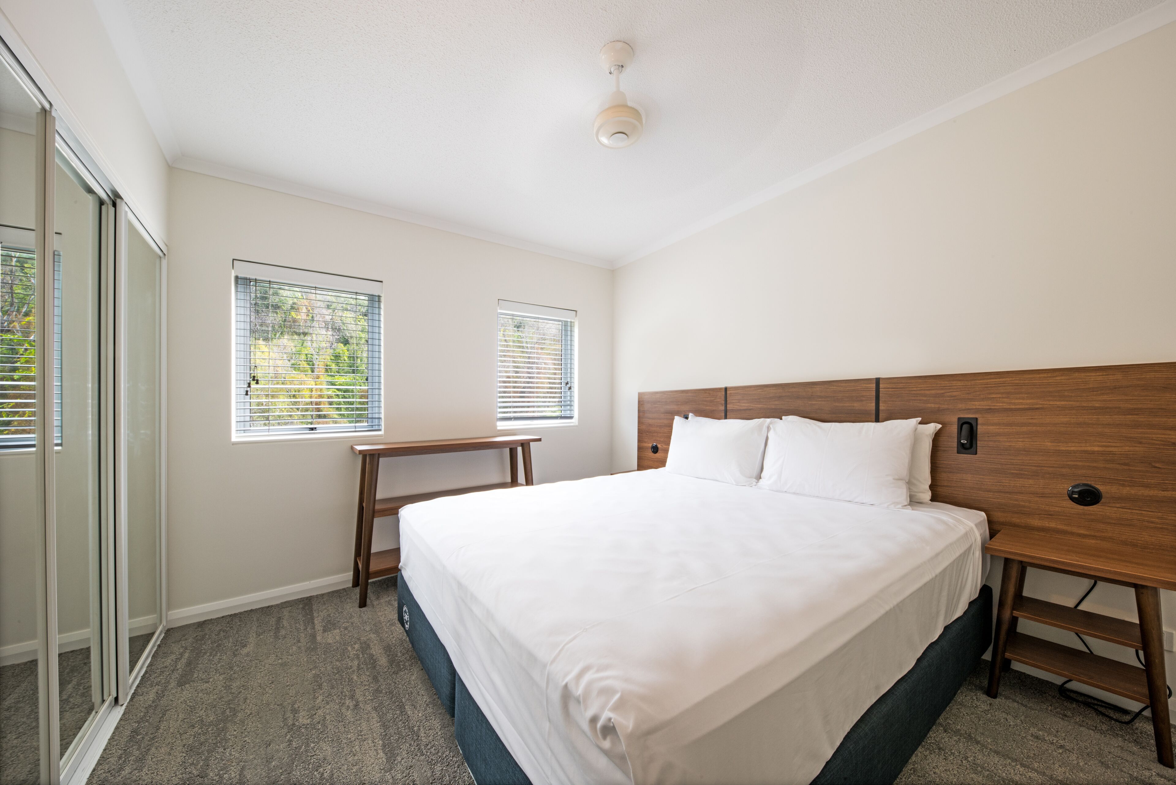 A bedroom with a large bed, a wooden headboard, a small table, and two windows with blinds.