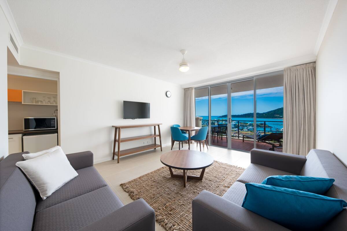 Una sala de estar moderna con sofá, una mesa de centro y televisor. Hay un balcón con vistas al mar.
