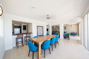 A spacious dining area with a wooden table and blue chairs, a kitchen in the background, and a view through a glass door.