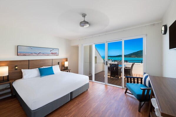 A hotel room with a large bed, a desk, and a balcony with a view of the sea.