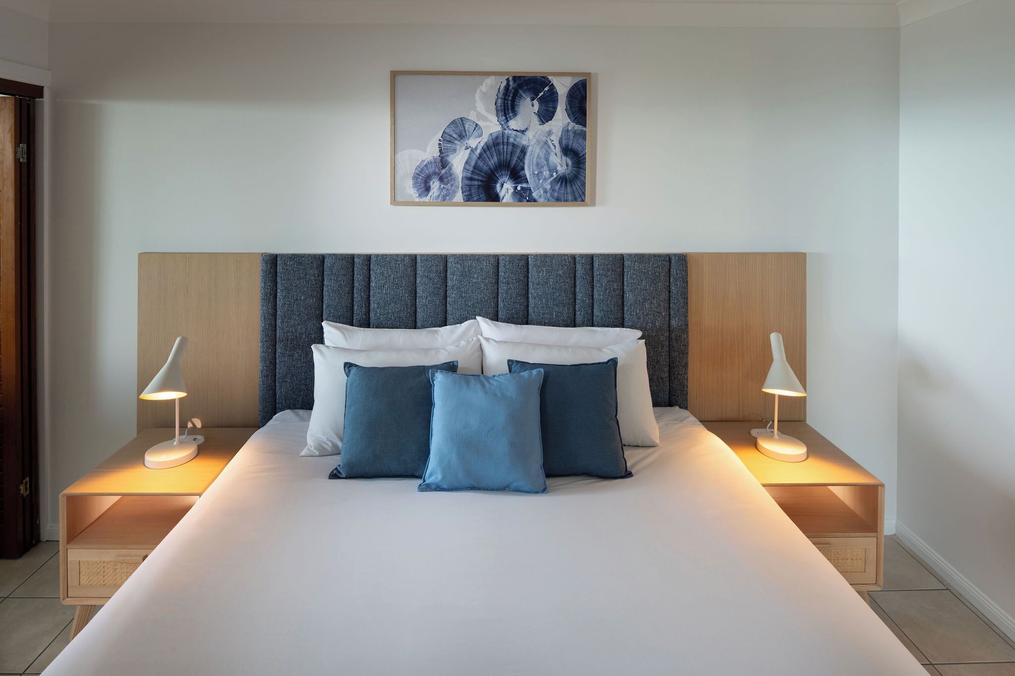 A neatly made bed with blue and white pillows, two bedside lamps, and a framed artwork above the bed.