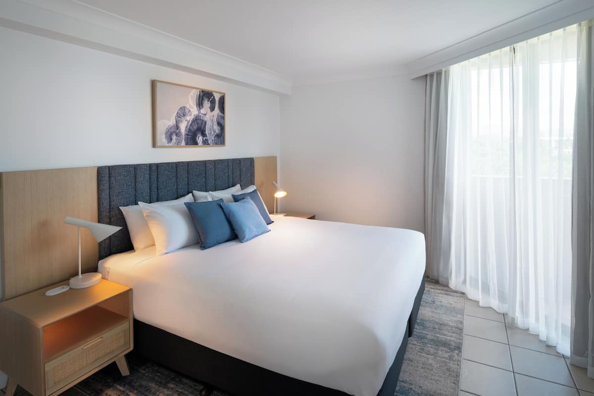 A neatly made bed with a headboard, a bedside table with a lamp, and a window with sheer curtains.