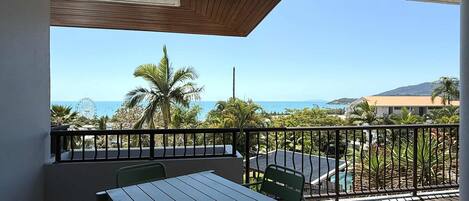 Un balcón con mesa y sillas, vista al mar, palmeras y una noria en la distancia.