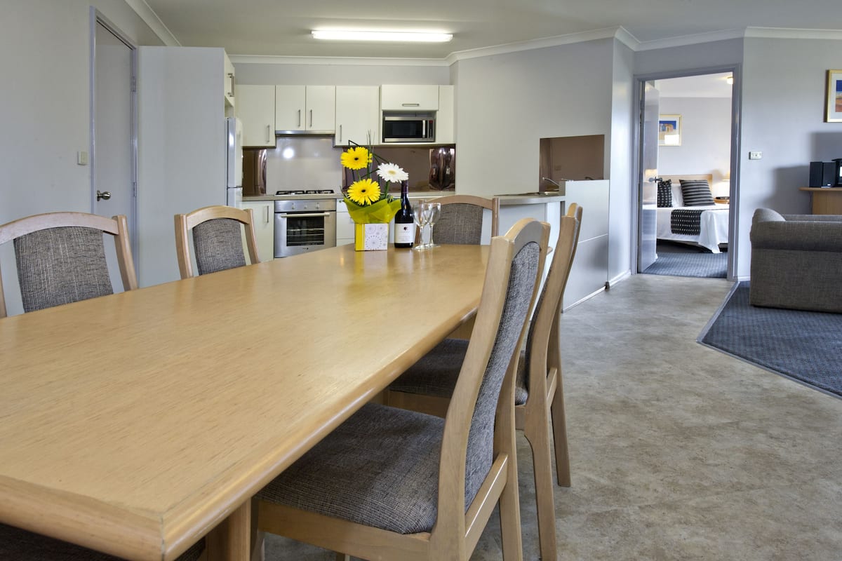 Un comedor con una mesa y sillas de madera, un florero con flores y una cocina al fondo.