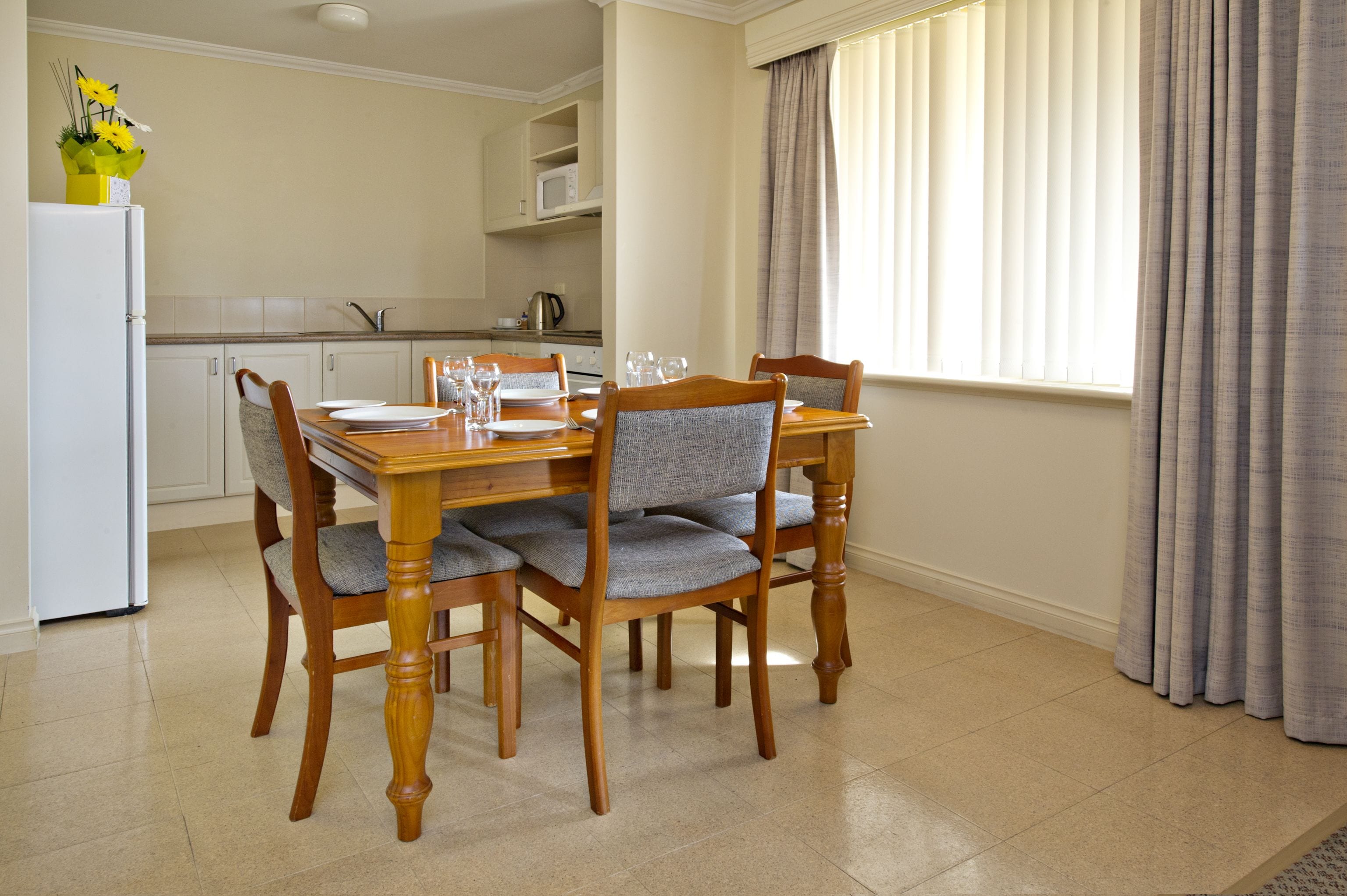 A dining area with a wooden table and chairs, a white refrigerator, and a window with curtains.