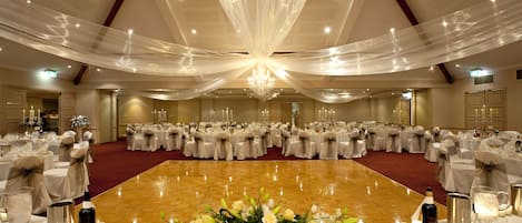 A banquet hall with round tables set for a formal event, featuring chandeliers and floral centerpieces.