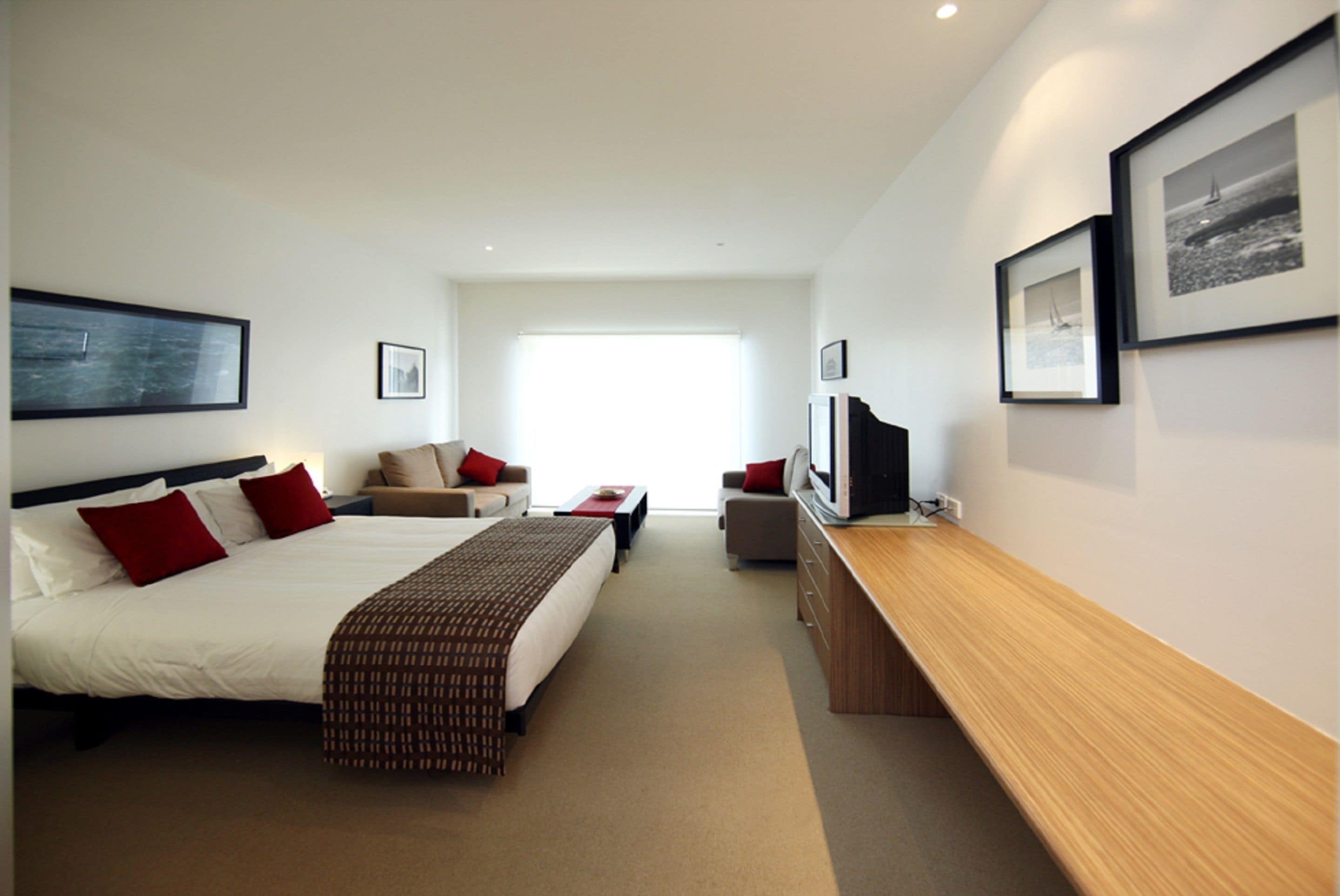 A hotel room with a large bed, a desk, a chair, and a TV. There are two windows and a door.