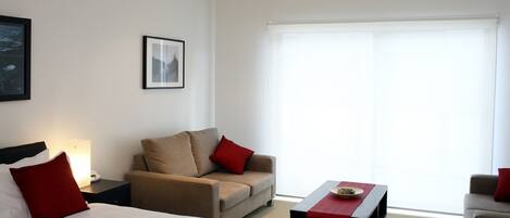 Quarto de hotel com cama, sofá, mesa de centro e janela com persianas.