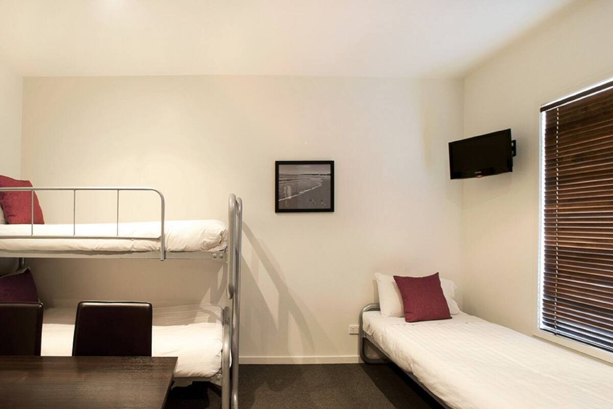 A dormitory room with bunk beds, a desk, a chair, a TV, and a window with blinds.