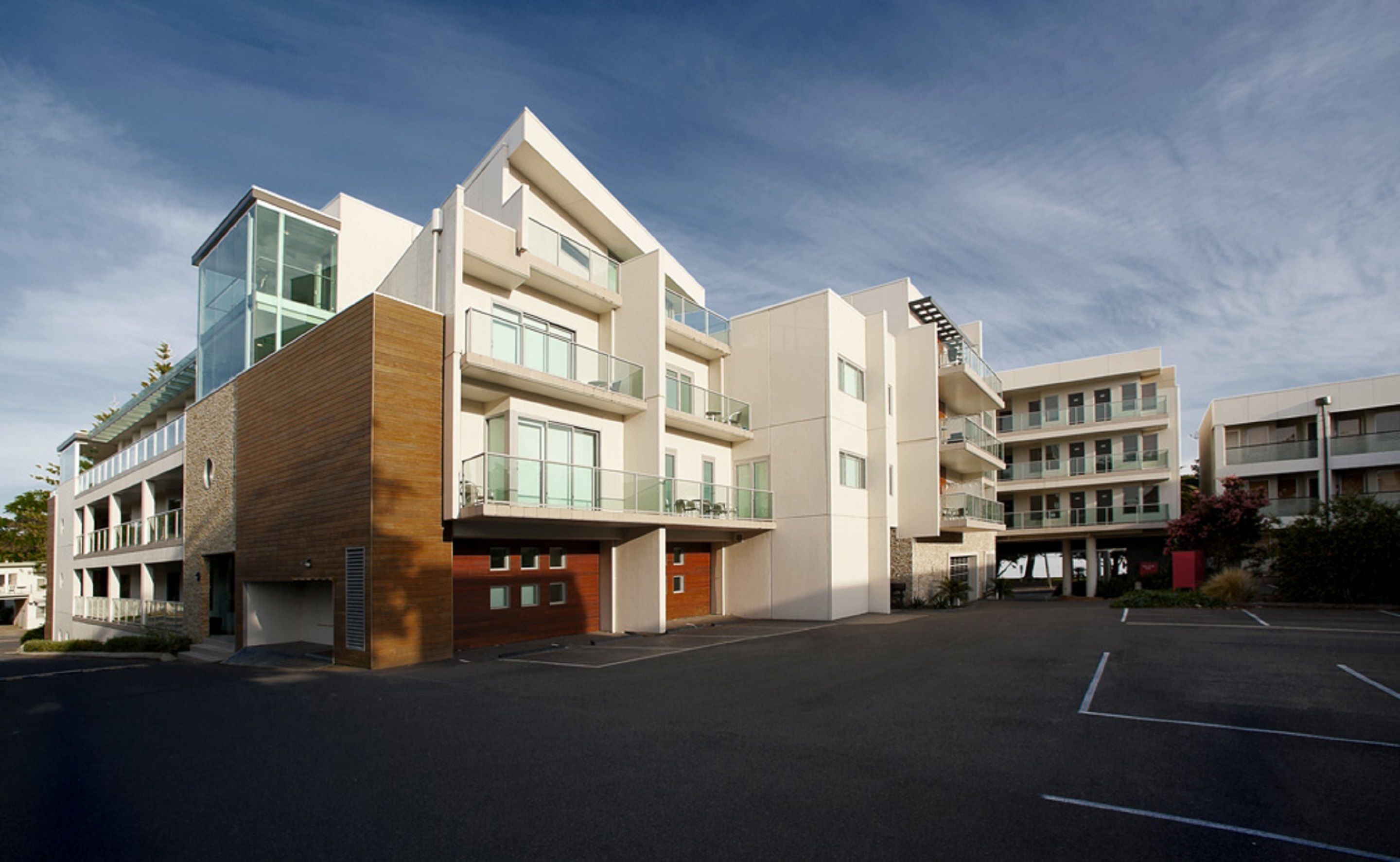 Un edificio moderno de varios pisos con fachadas de vidrio y madera, varios balcones y un estacionamiento.