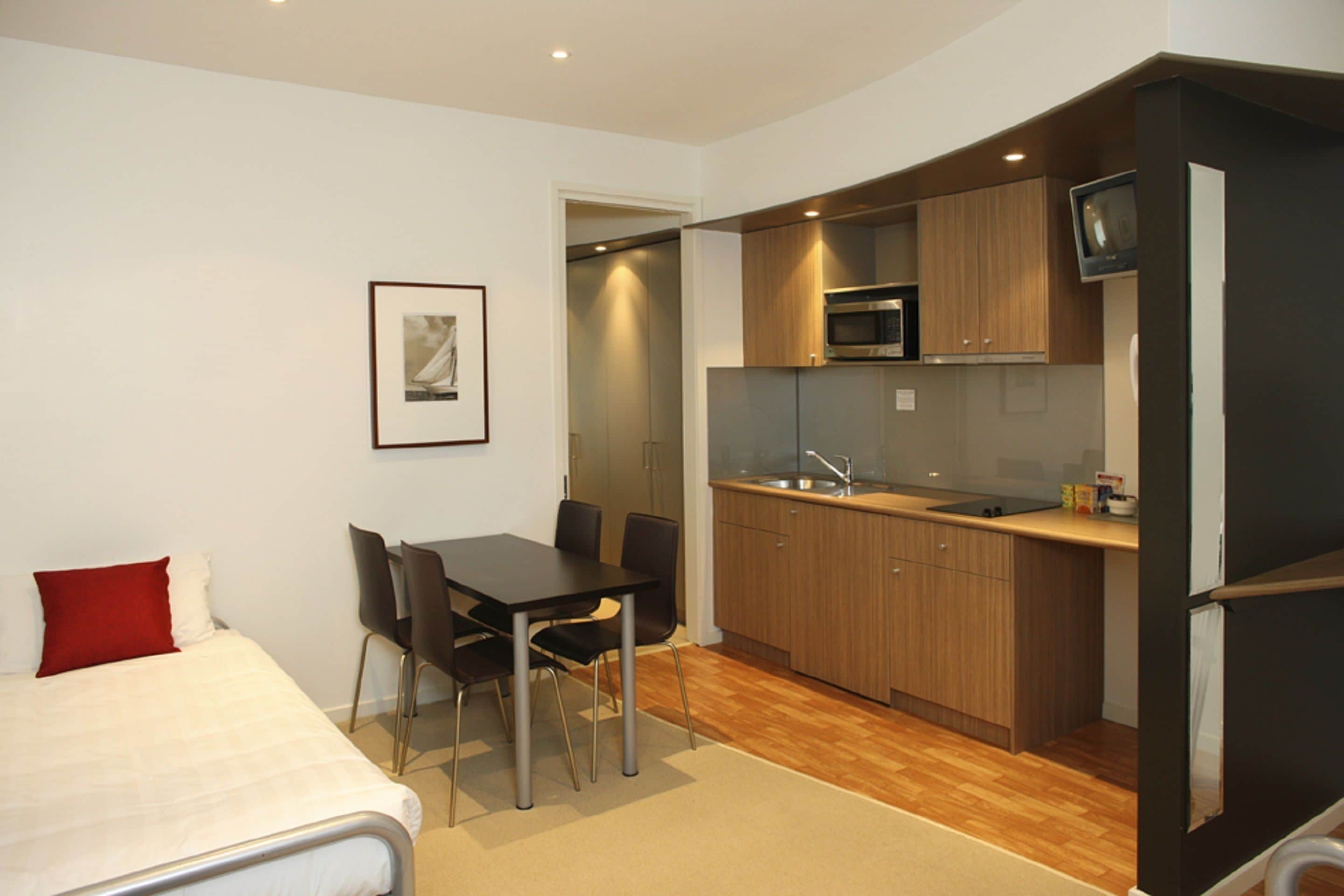 A compact hotel room with a bed, a dining table with chairs, a kitchenette with a microwave, and a TV.