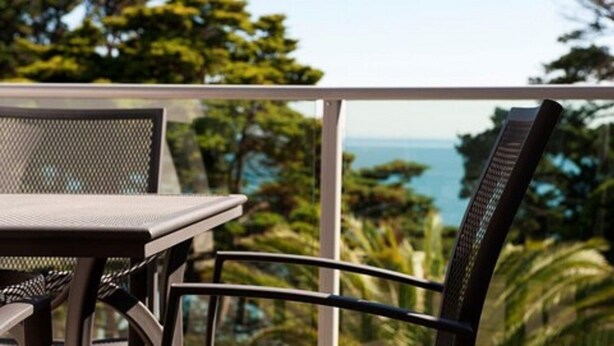 Un balcón con mesa y sillas, con vista a un paisaje tropical con árboles y el mar.