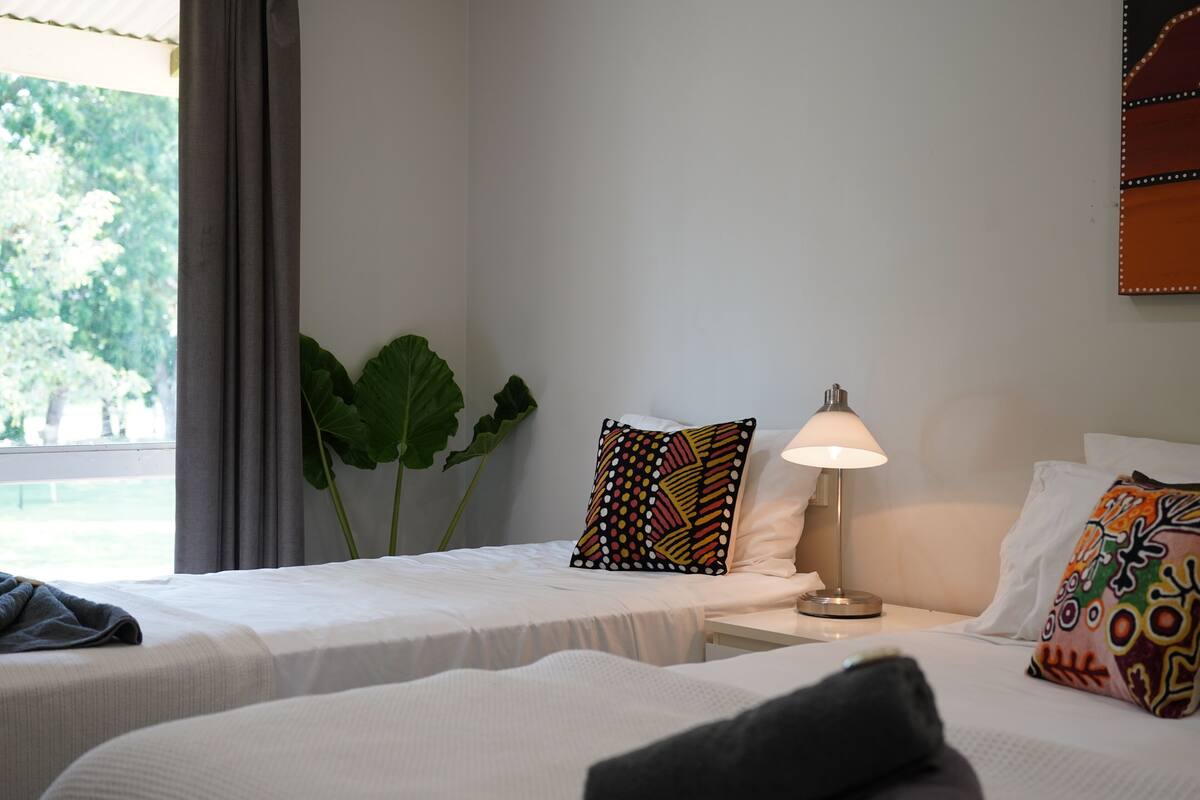 Habitación de hotel con dos camas, una mesita de noche con lámpara, una planta grande y una ventana con vistas a árboles.