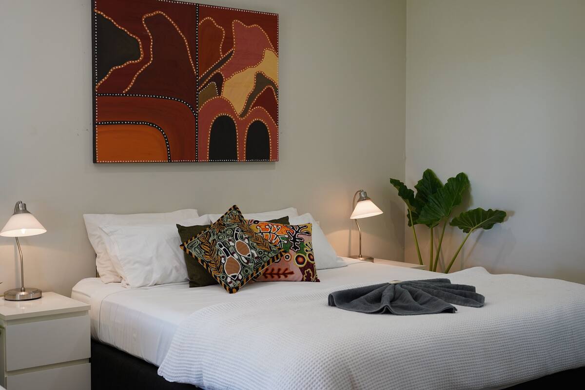 Habitación de hotel con una cama bien hecha, dos lámparas de noche, un cuadro abstracto grande y una planta en maceta.