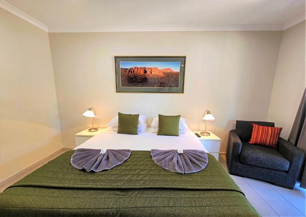 Habitación de hotel con una cama, dos mesitas de noche con lámparas, un cuadro enmarcado en la pared y un sillón gris.