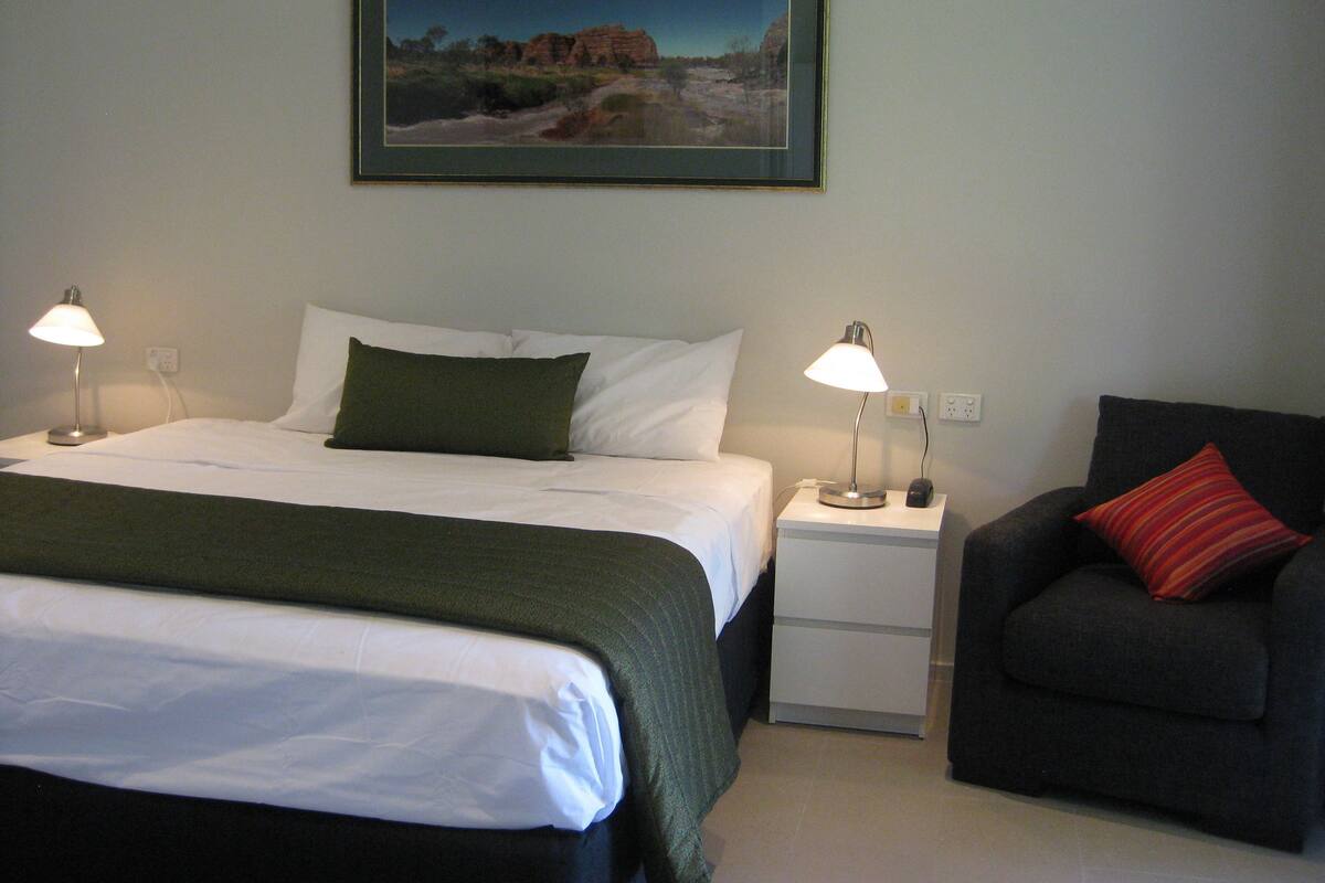 Habitación de hotel con cama, dos lámparas de noche, mesita de noche, una silla e imagen enmarcada en la pared.