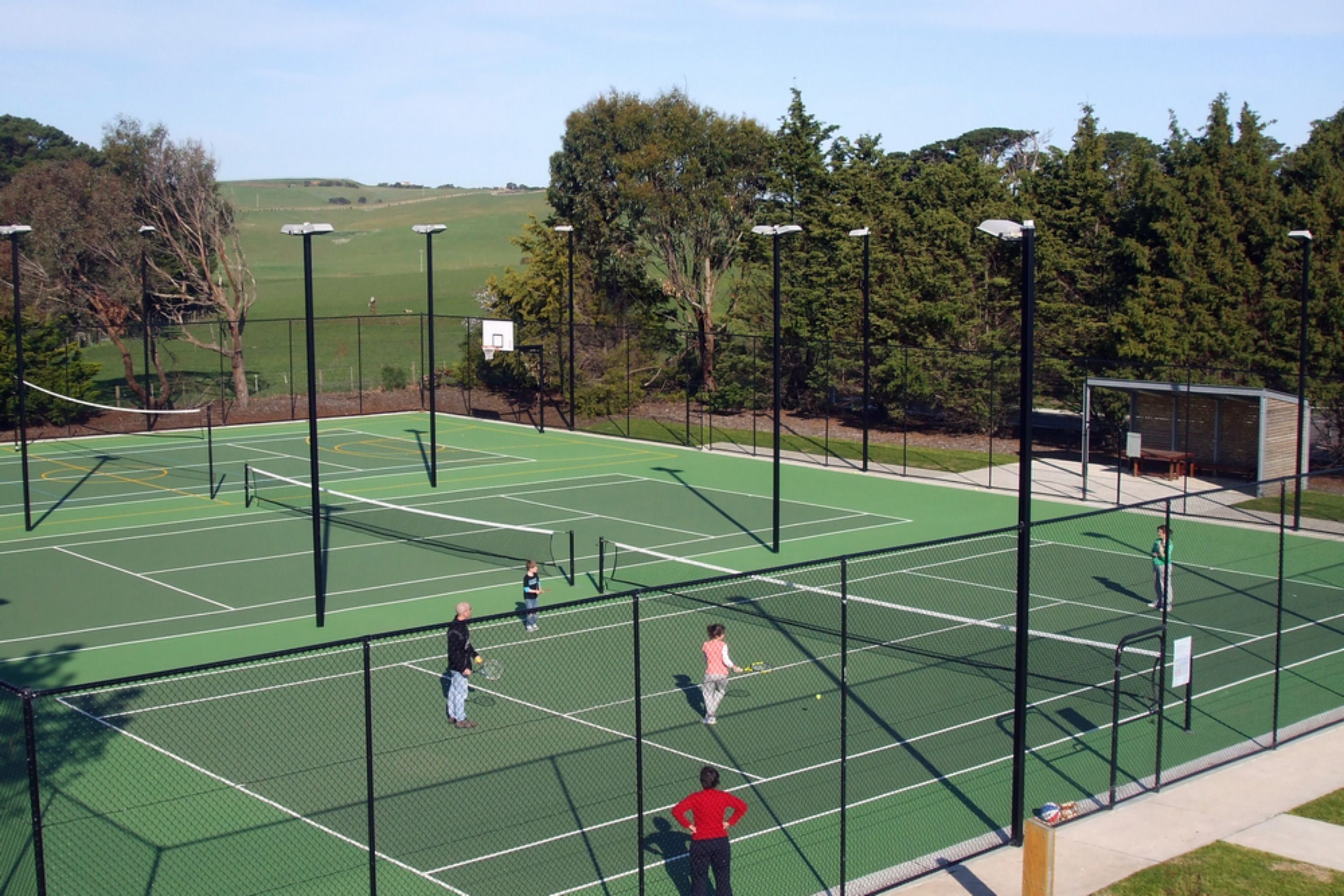 An outdoor tennis court with multiple players, surrounded by trees and a fence.