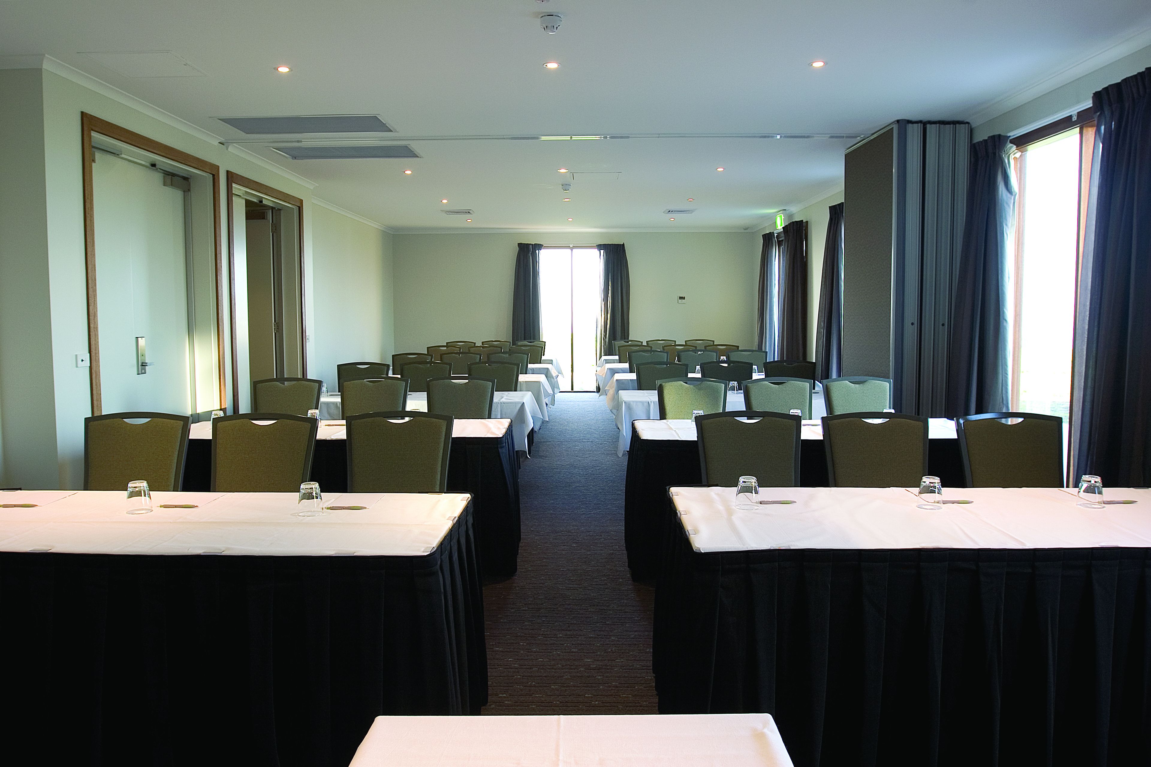 A conference room with rows of tables and chairs, large windows with curtains, and a carpeted floor.