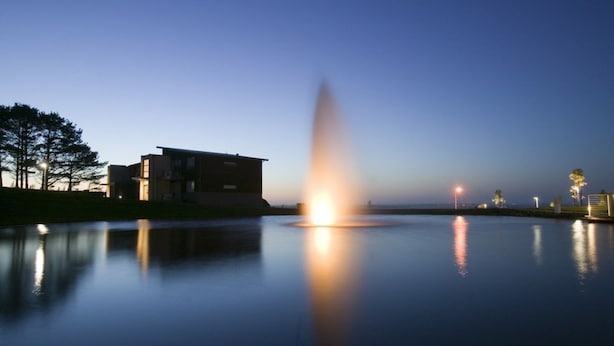 A calm body of water reflecting a building and a fountain.