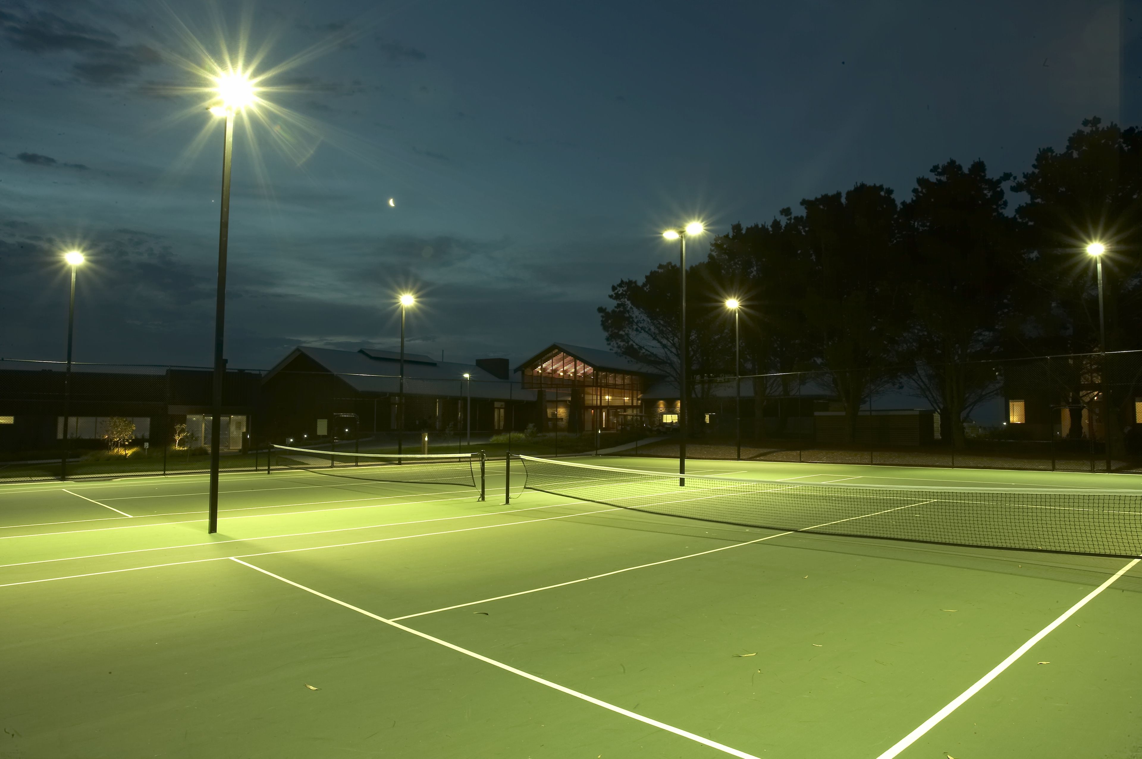 Cancha de tenis exterior iluminada con múltiples focos durante la noche.
