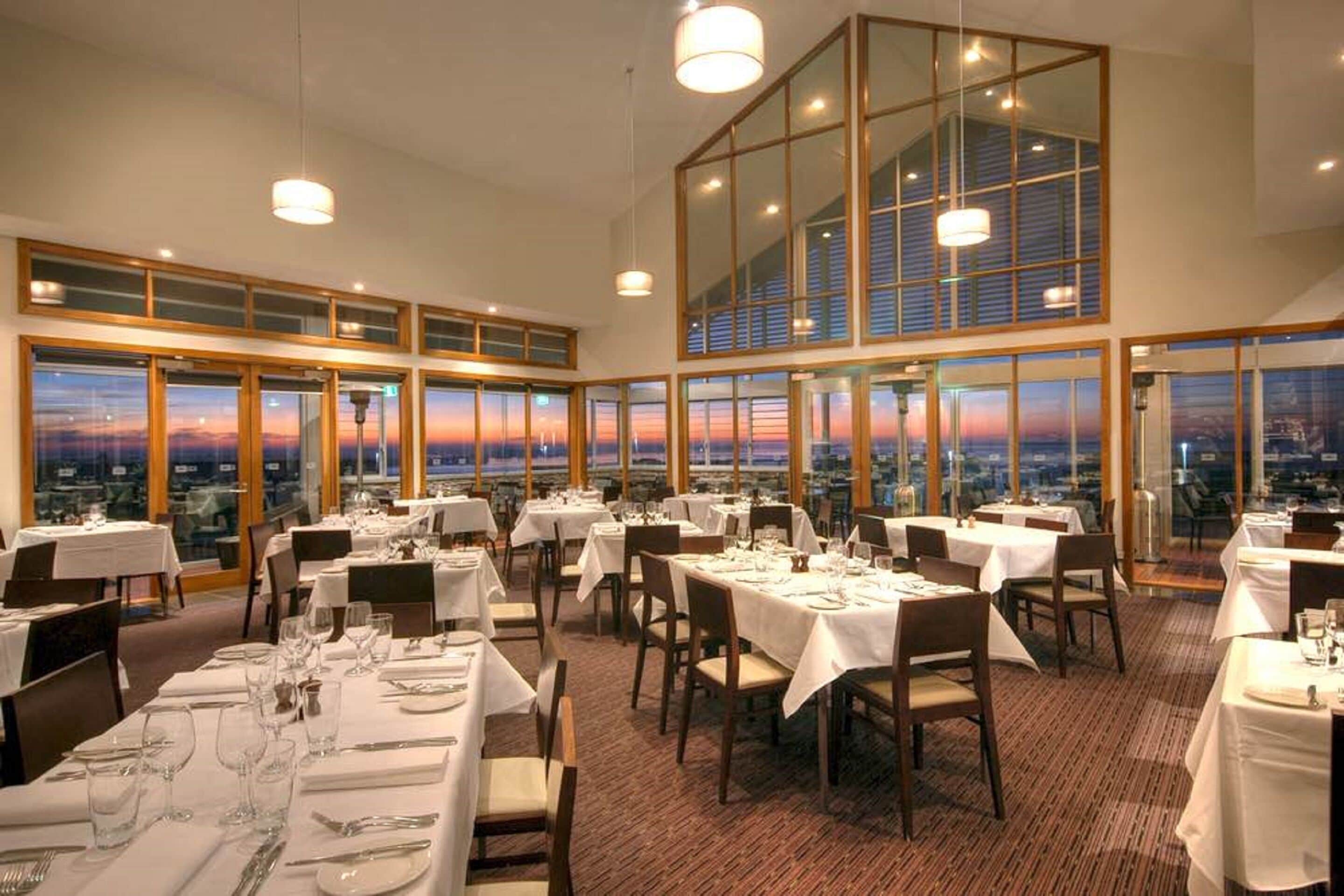 Un restaurant amb diverses taules preparades per a dinar, grans finestres que ofereixen una vista de la ciutat i un sostre alt amb llums penjants.