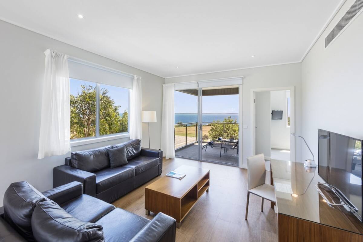 Una sala de estar moderna con sofá, mesa de centro y televisor. Cuenta con ventanas amplias que ofrecen vistas al exterior.