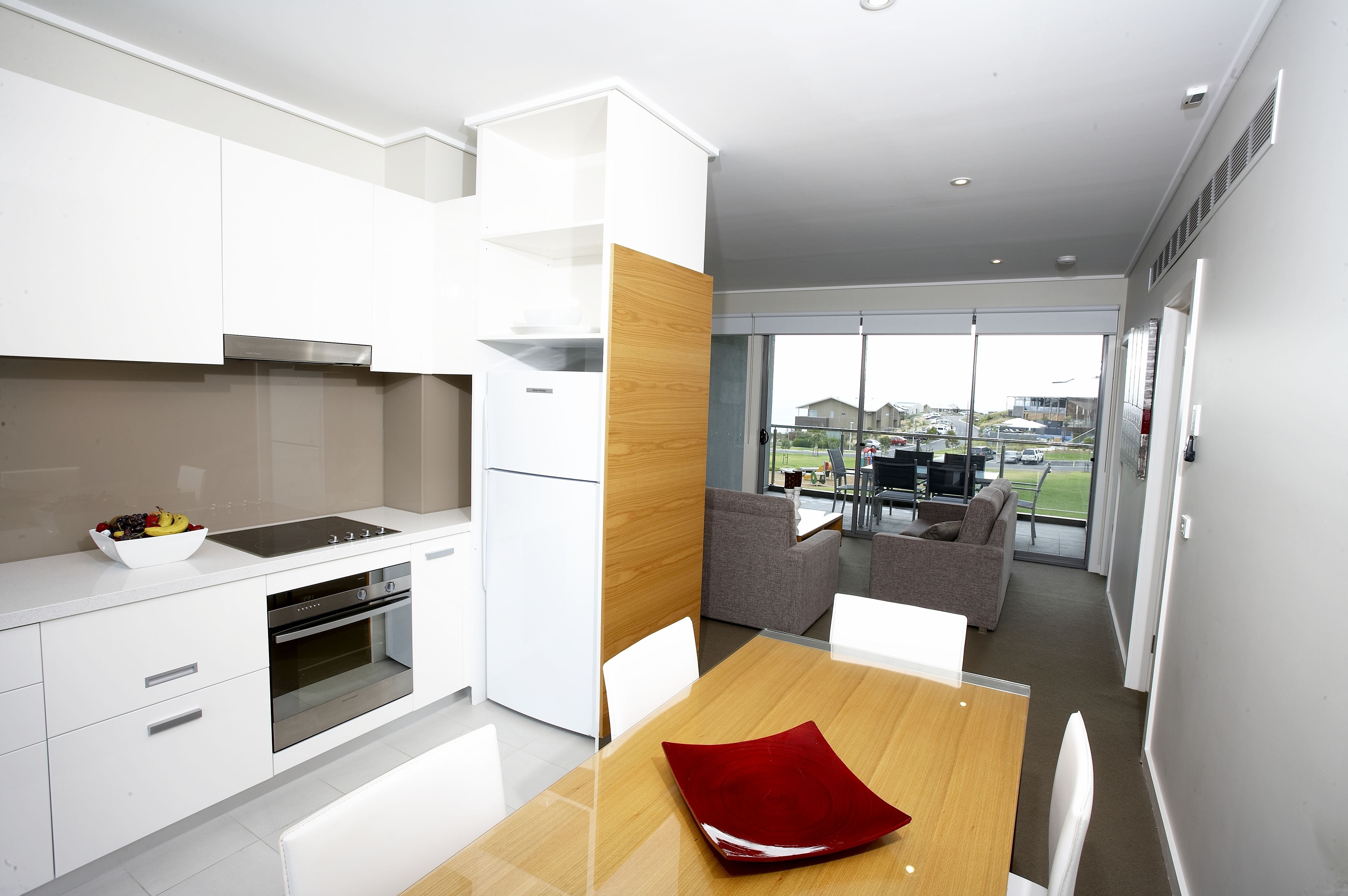 Una cocina moderna con armarios blancos, un muro decorativo de madera y un comedor con un cojín rojo.