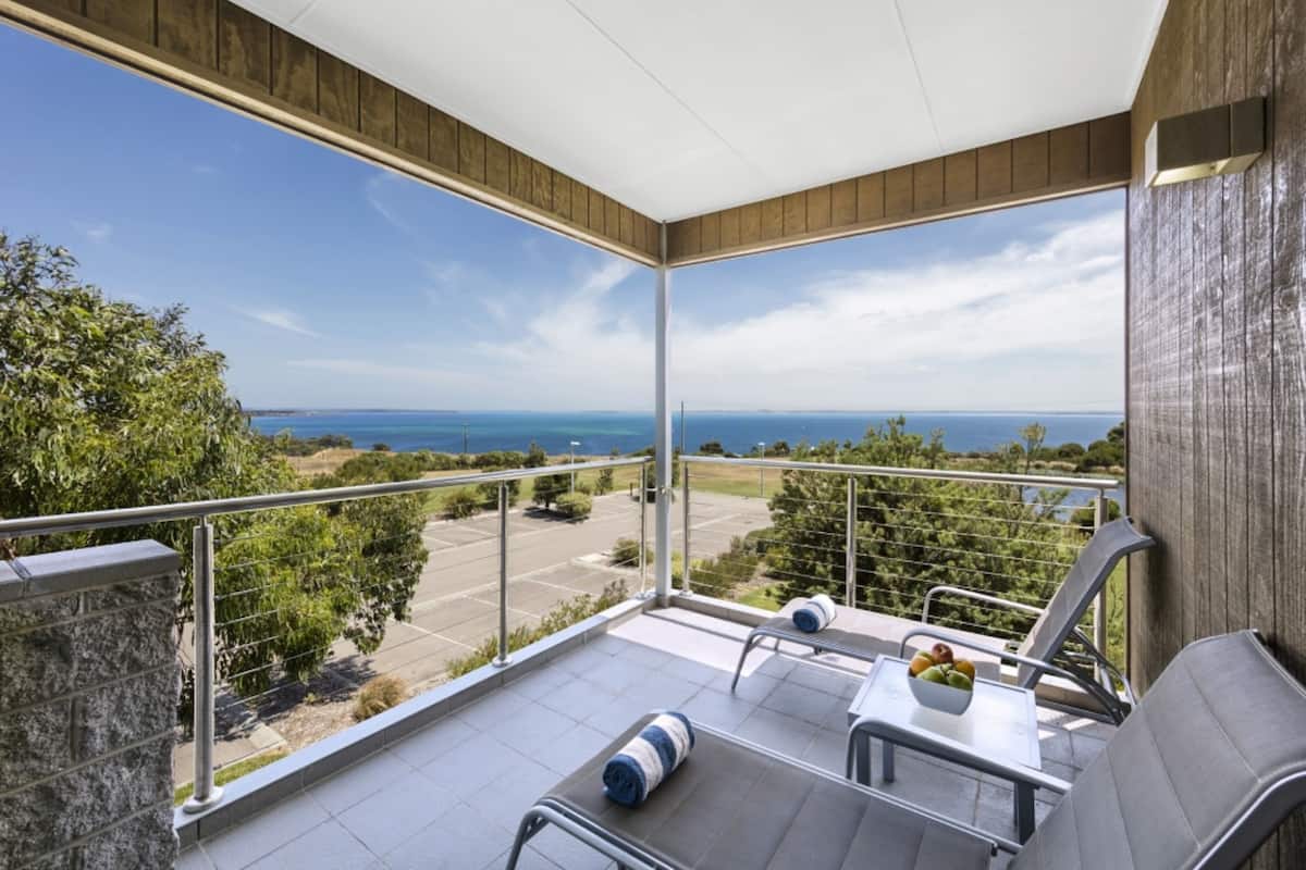 Un balcón con vista al mar, una mesa con fruta y dos sillones de descanso.