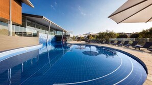 A modern swimming pool with a tiled edge, surrounded by lounge chairs and umbrellas, and a building with large windows in the background.