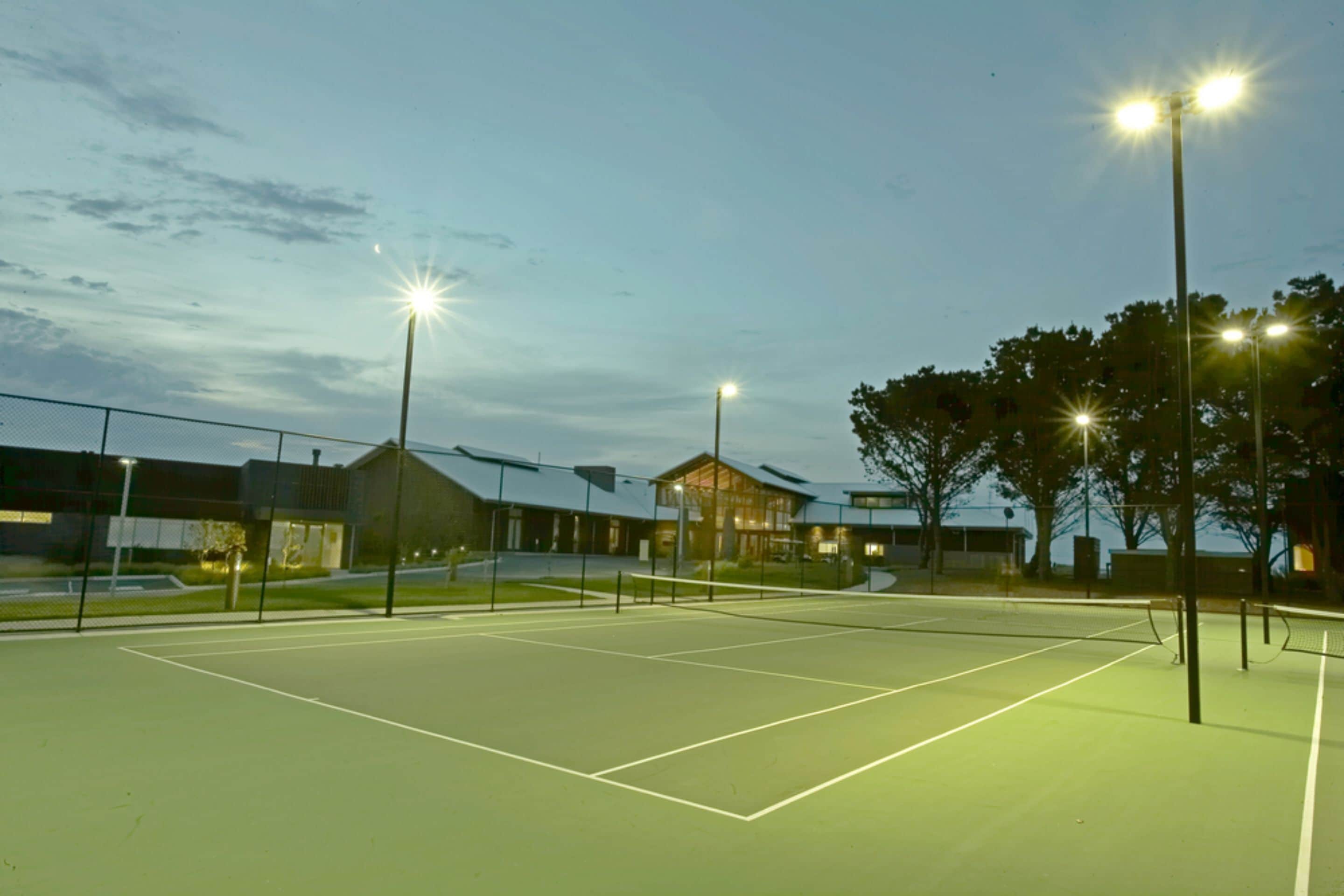 An outdoor tennis court at dusk with floodlights, surrounded by a fence and trees.
