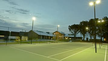 An outdoor tennis court at dusk with floodlights, surrounded by a fence and trees.