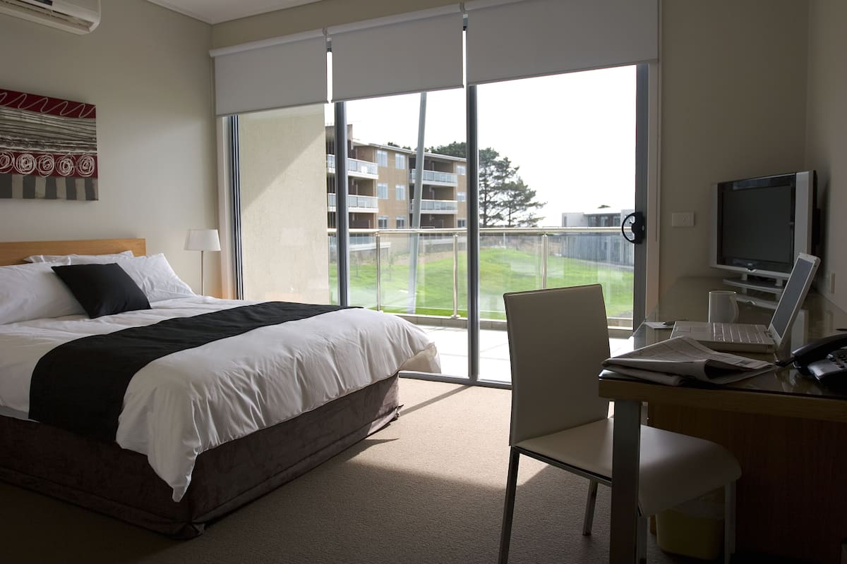 Habitación de hotel con una cama grande, un escritorio con una computadora portátil, un televisor y vistas a un edificio y un árbol a través de ventanas amplias.
