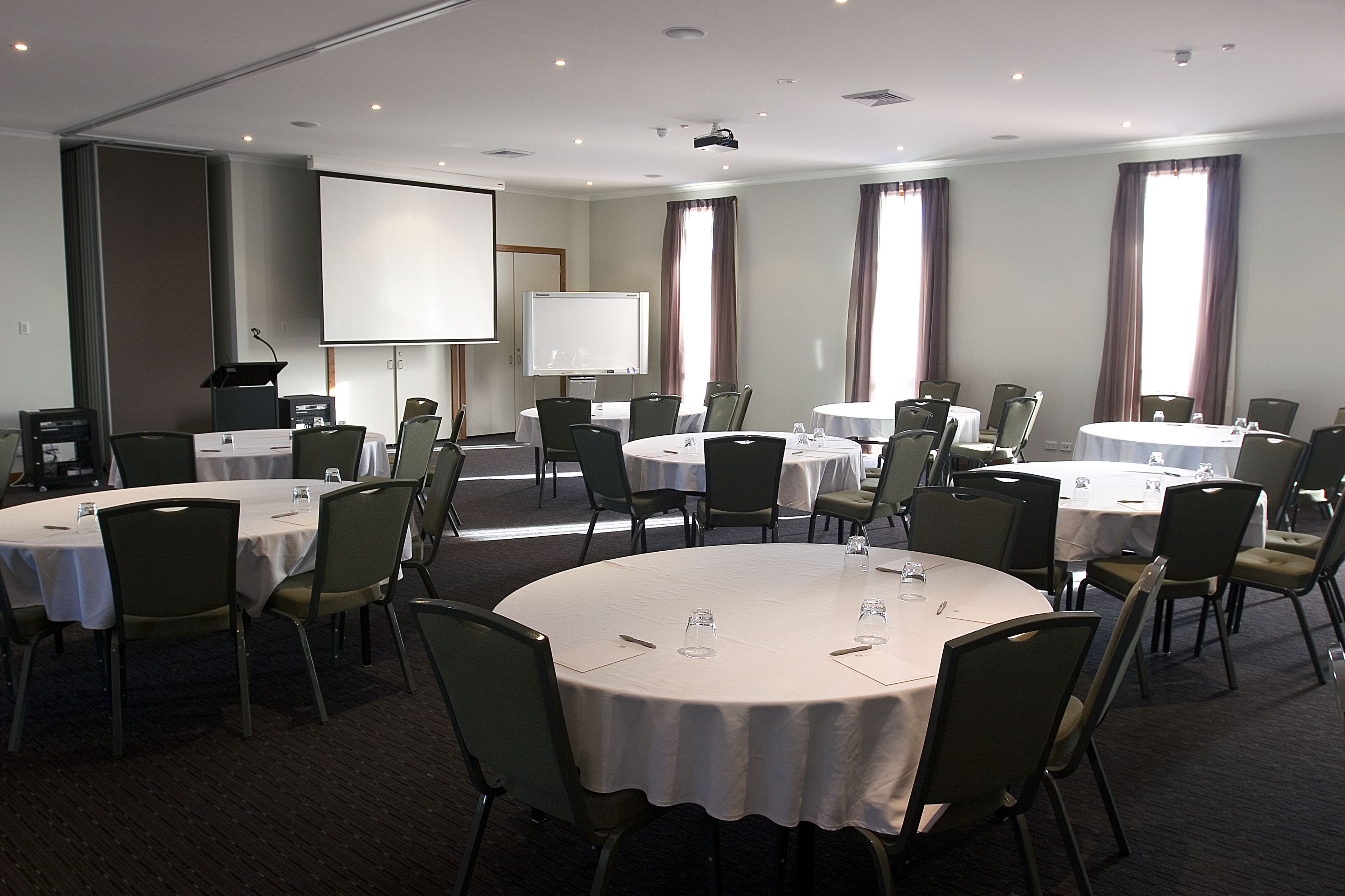 Un salón de conferencias con varias mesas redondas, sillas, una pantalla de proyector y una pizarra.