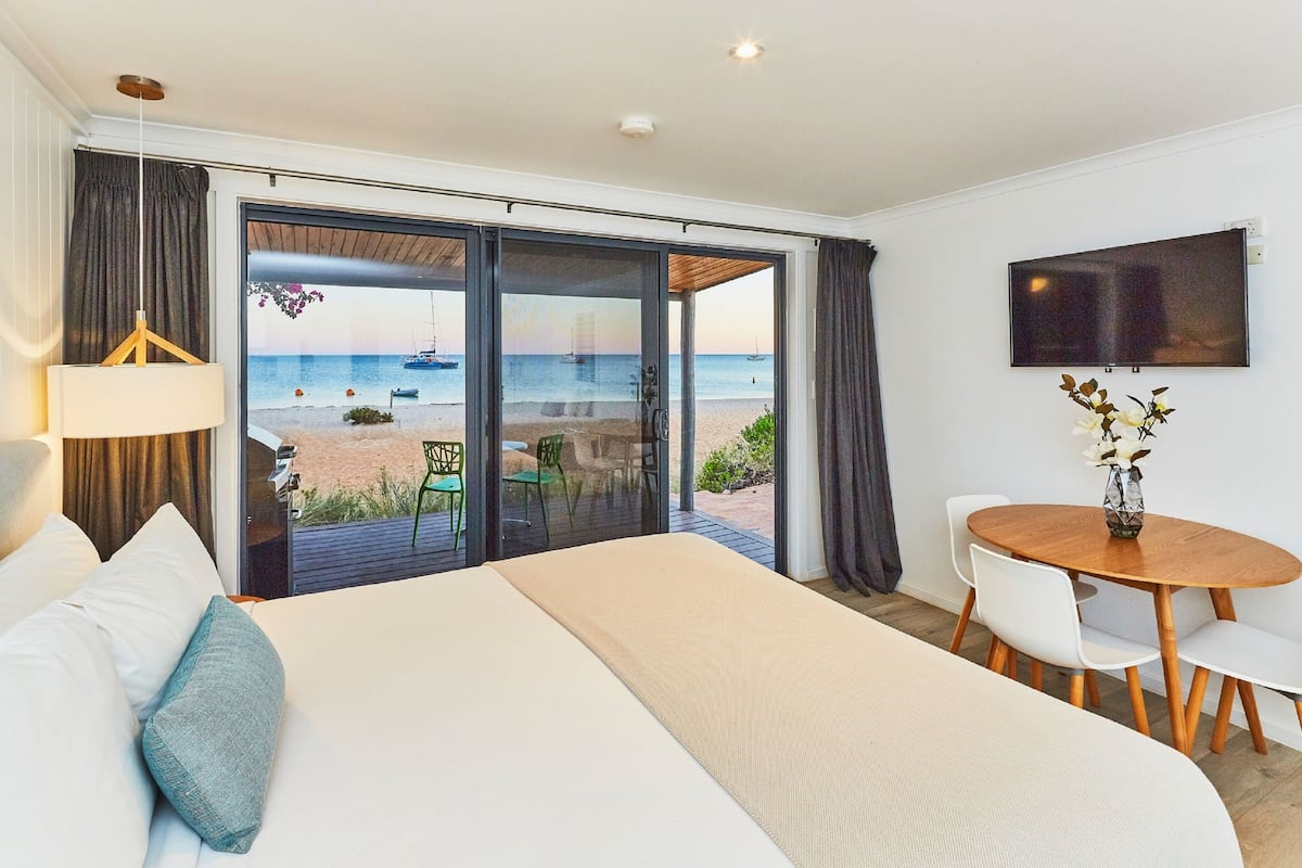 Habitación de hotel con una cama grande, una mesa de comedor con sillas, un televisor y vista a la playa y al océano a través de puertas correderas de vidrio.