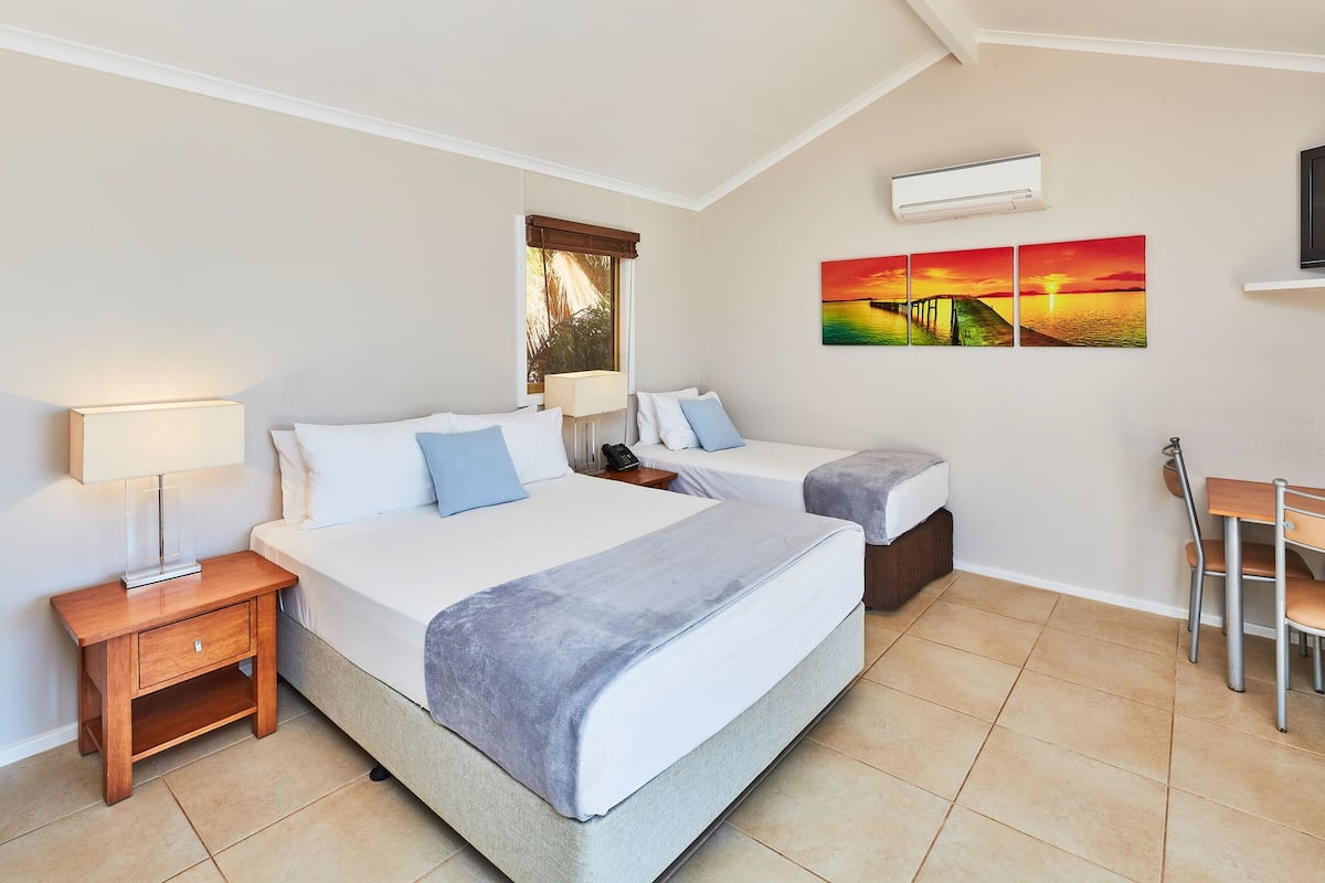 Un dormitorio con dos camas, un escritorio y una silla. Cuenta con una ventana con persianas, un equipo de aire acondicionado y un cuadro en la pared.