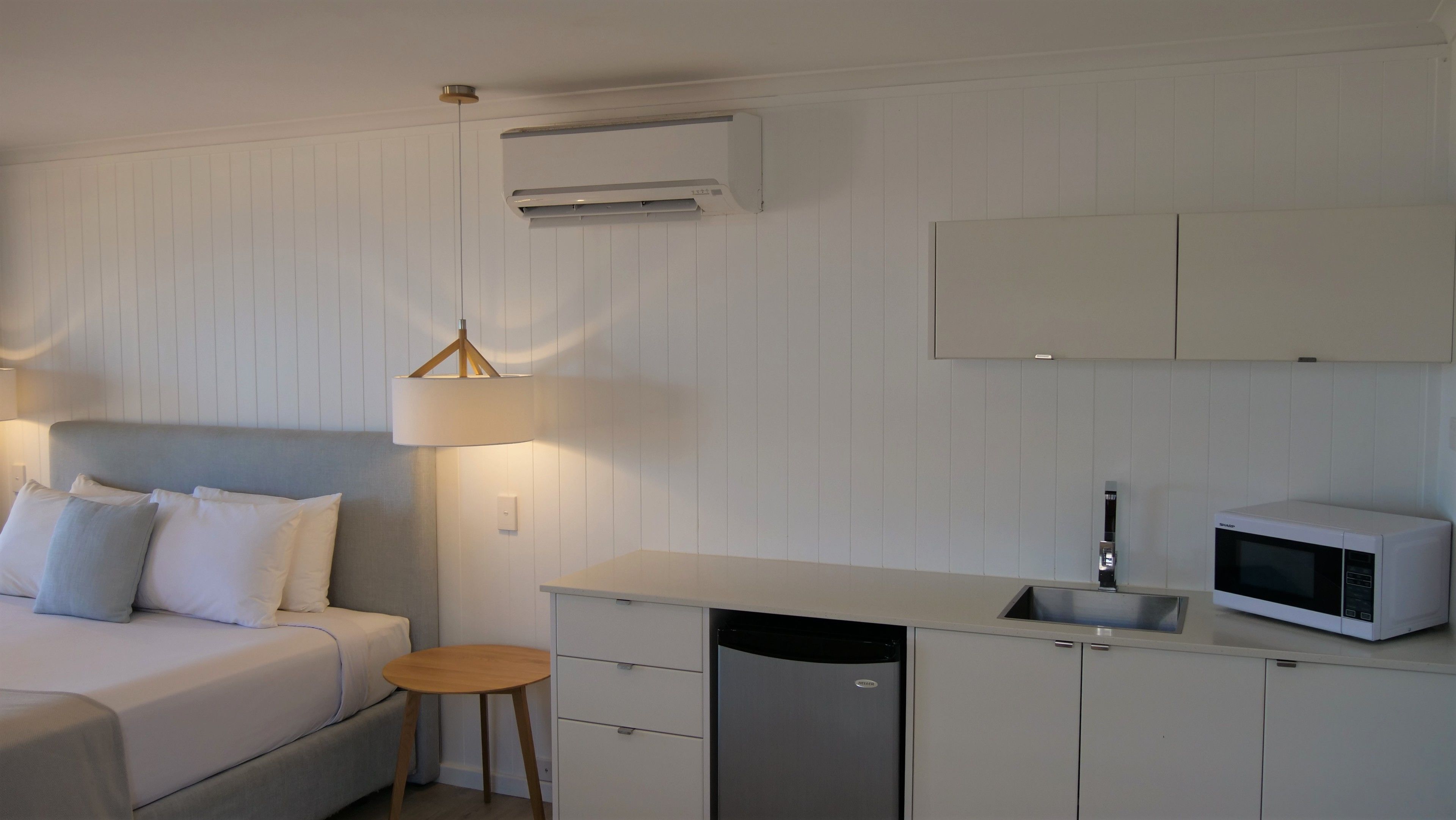 Une chambre compacte et moderne, équipée d’un lit, d’une lampe de chevet, d’une kitchenette et d’un système de climatisation intégré.