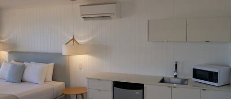 Une chambre compacte et moderne, équipée d’un lit, d’une lampe de chevet, d’une kitchenette et d’un système de climatisation intégré.