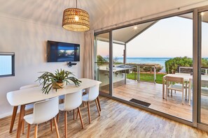A modern interior with a dining area, a TV, and a large sliding glass door leading to an outdoor deck with a view of the sea. - RAC Monkey Mia Dolphin Resort (Monkey Mia)