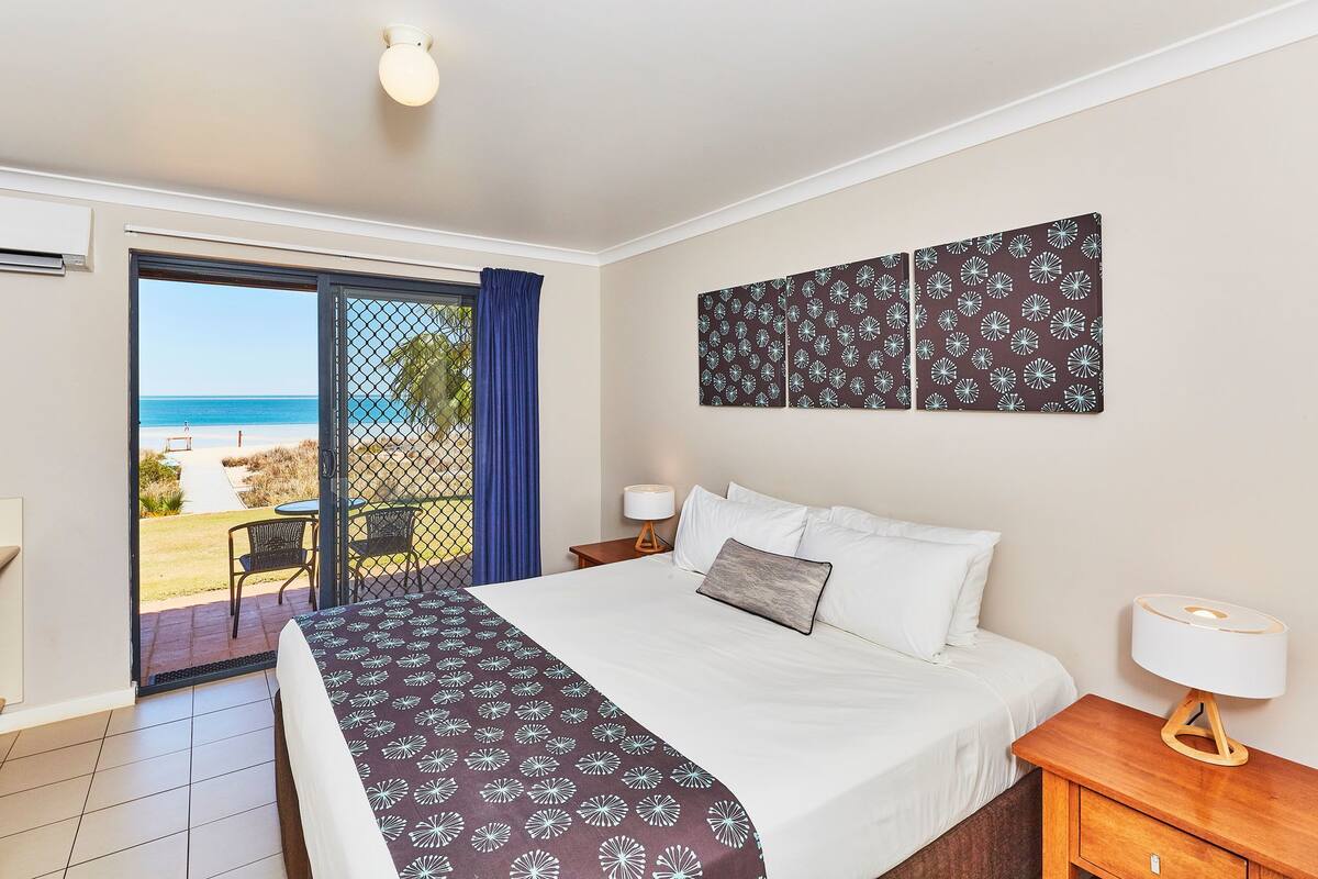 Habitación de hotel con cama, mesita de noche, lámpara, vista a la playa y puerta corrediza al balcón.