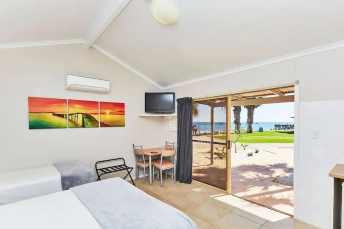 A hotel room with a bed, a desk with a chair, a TV, and a painting. There is a sliding glass door leading to a balcony with a view of the beach.