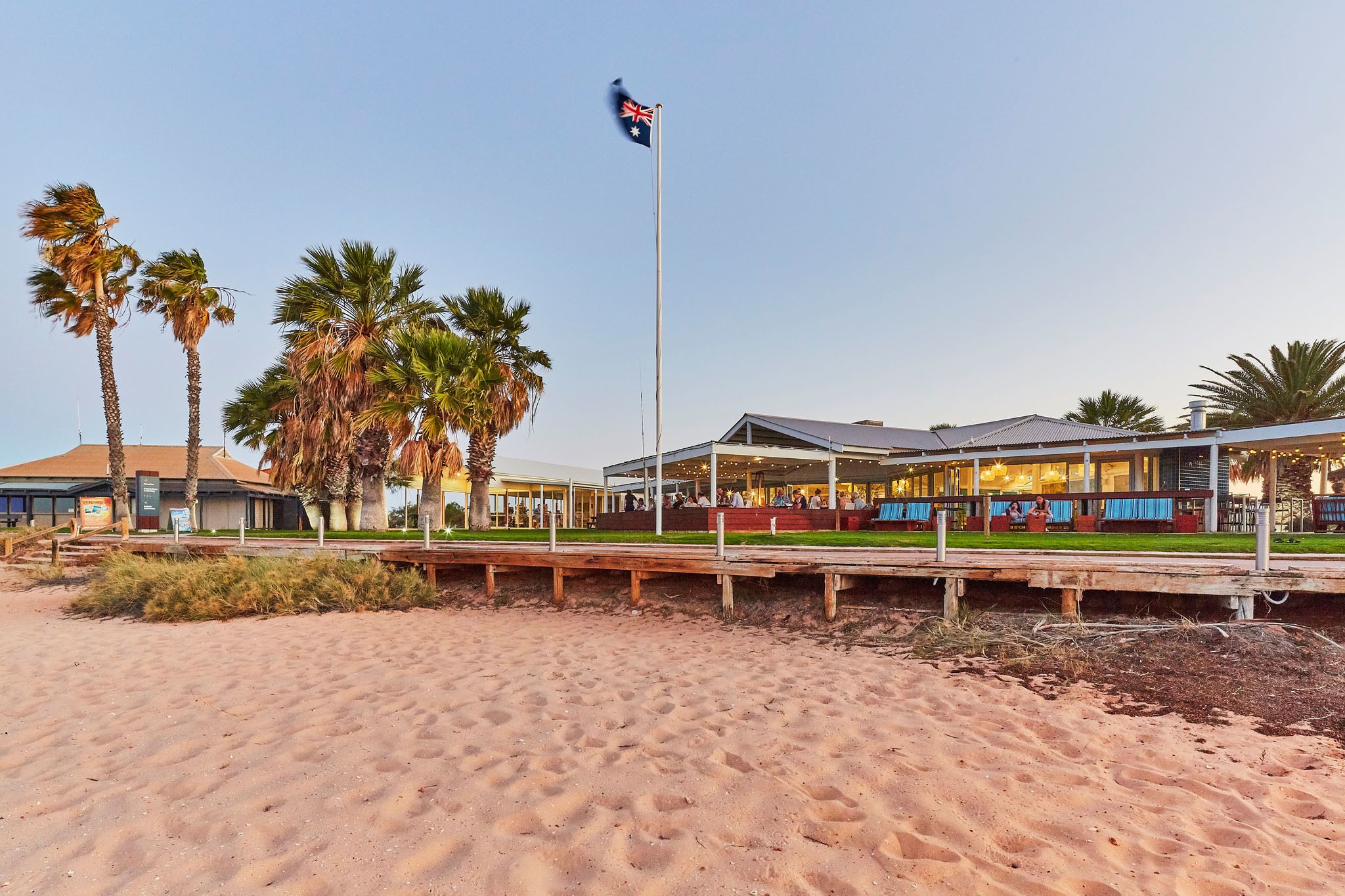 Un hotel frente a la playa con un asta de bandera, palmeras y una zona de estar al aire libre cubierta.