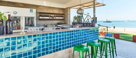 An outdoor bar with a blue-tiled counter, green stools, and a view of the sea.