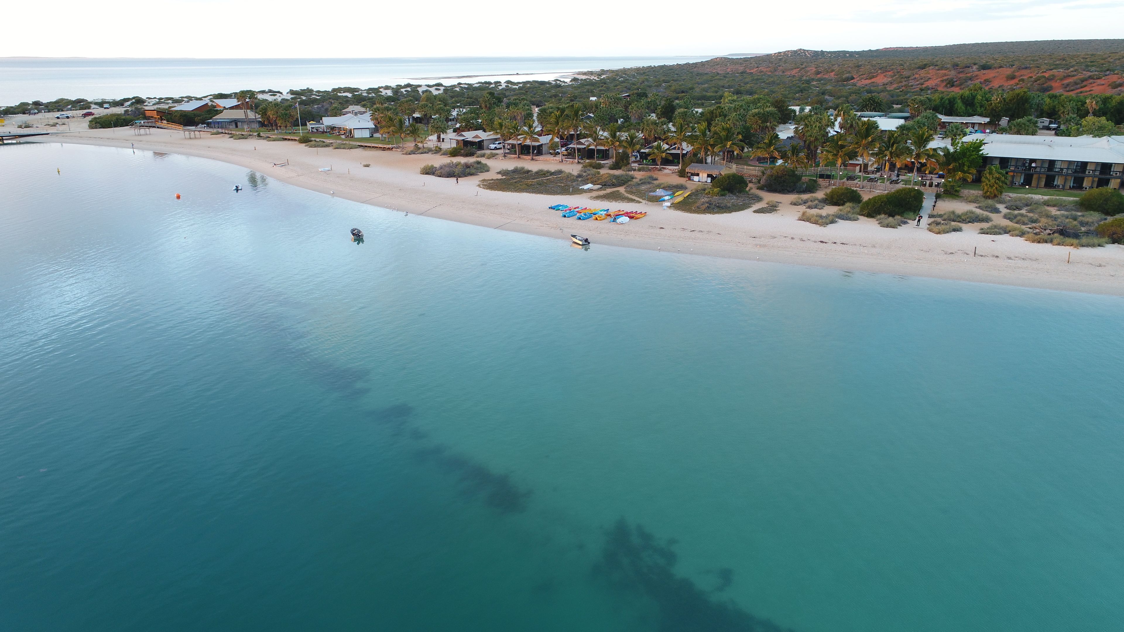 An aerial view of a coastal area with a beach, clear blue water, and a variety of buildings including a large resort with multiple structures and palm trees.