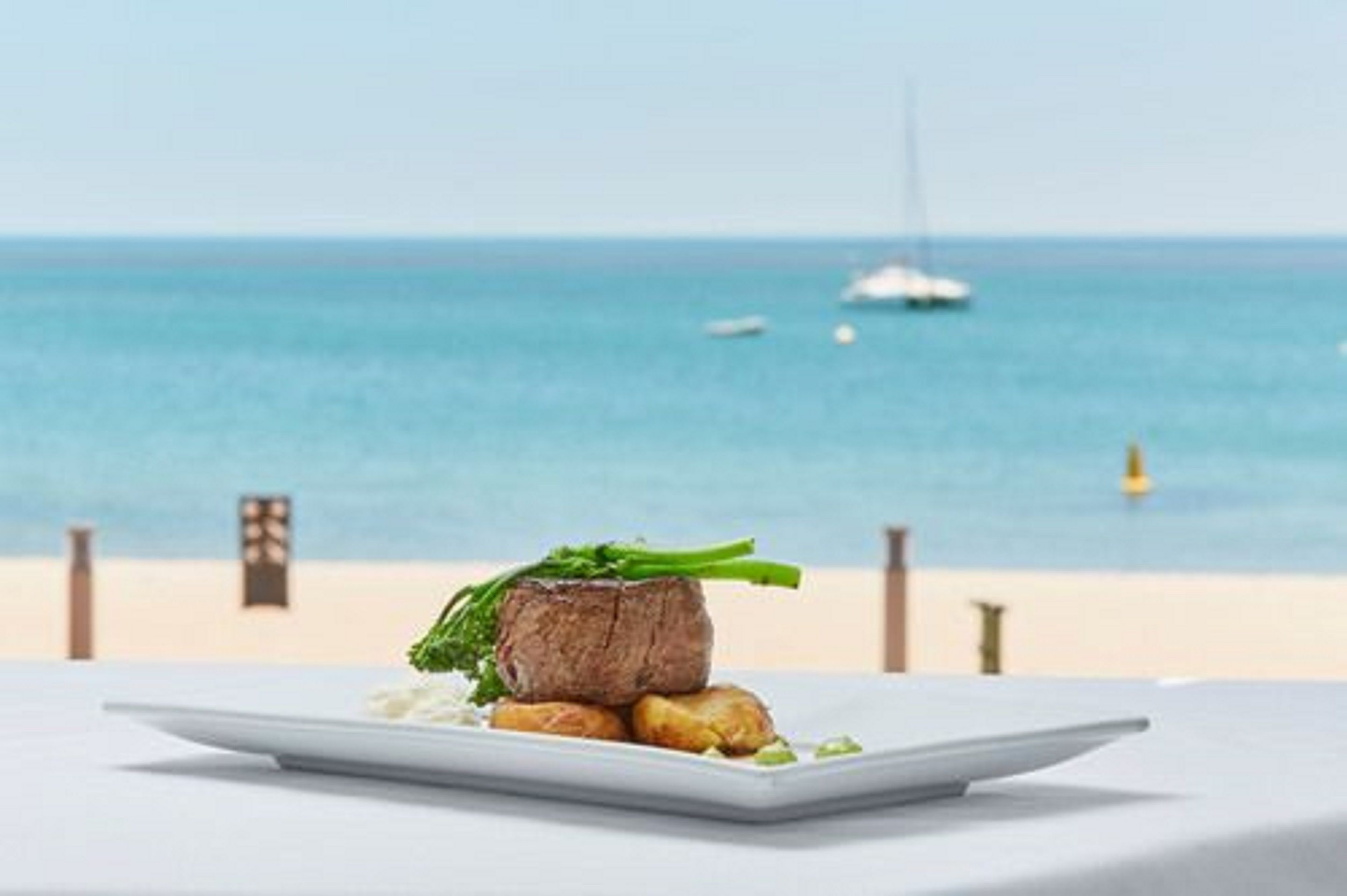 A plate of food with a steak and vegetables on a white table with a beach and ocean view in the background.