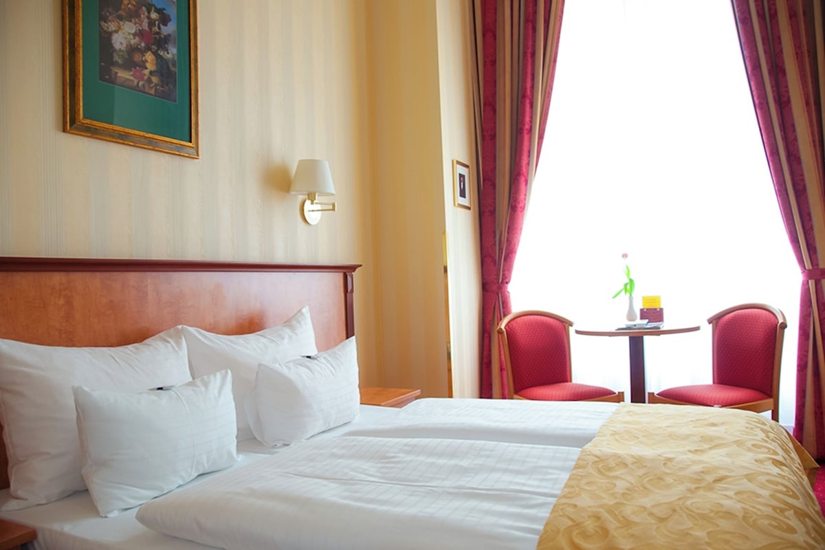 Una habitación de hotel con una cama grande, dos sillas rojas, una mesita y una ventana con cortinas.