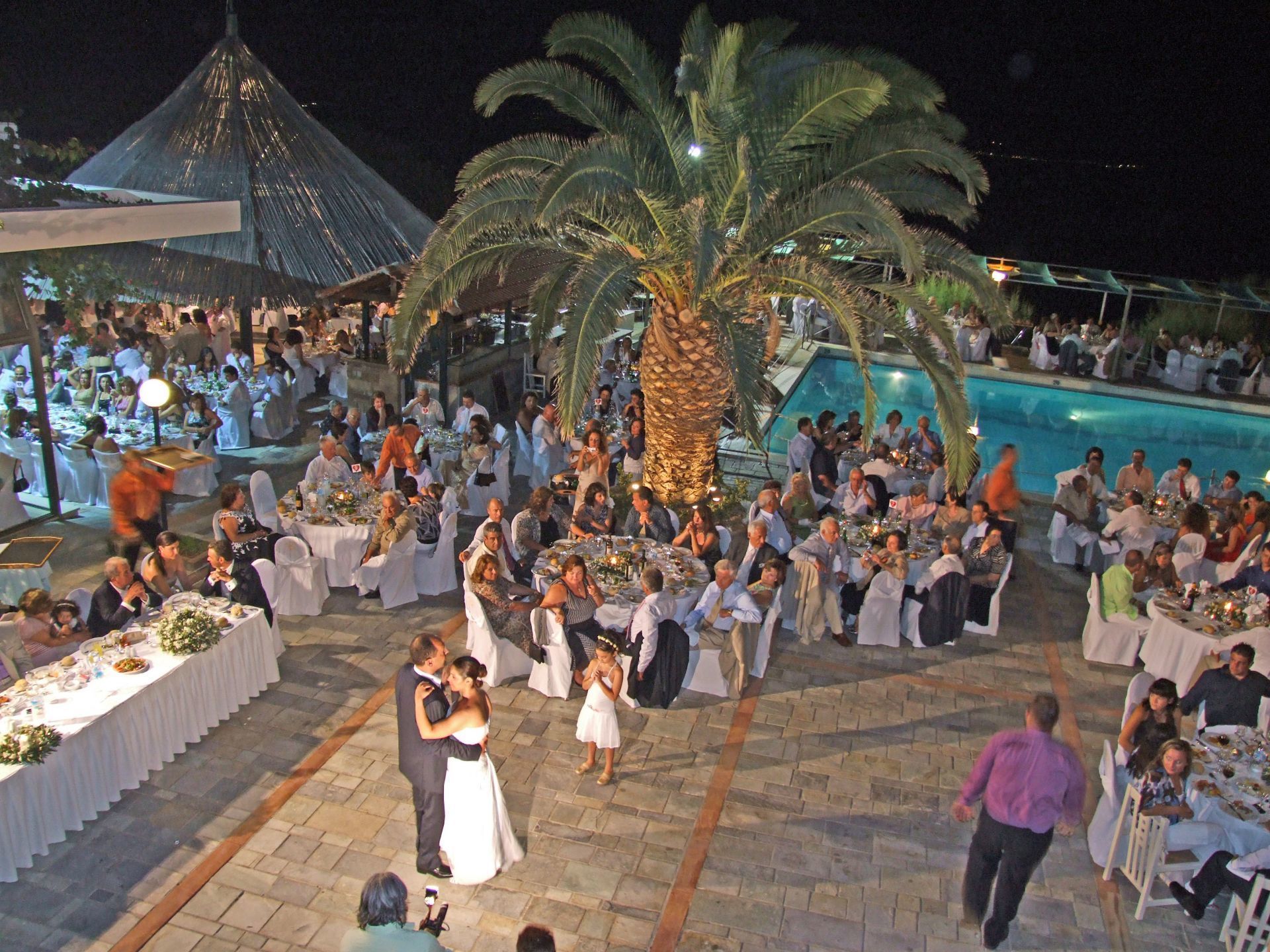 Una recepción de boda al aire libre con invitados sentados en mesas redondas, una pareja bailando en el centro y una gran palmera.