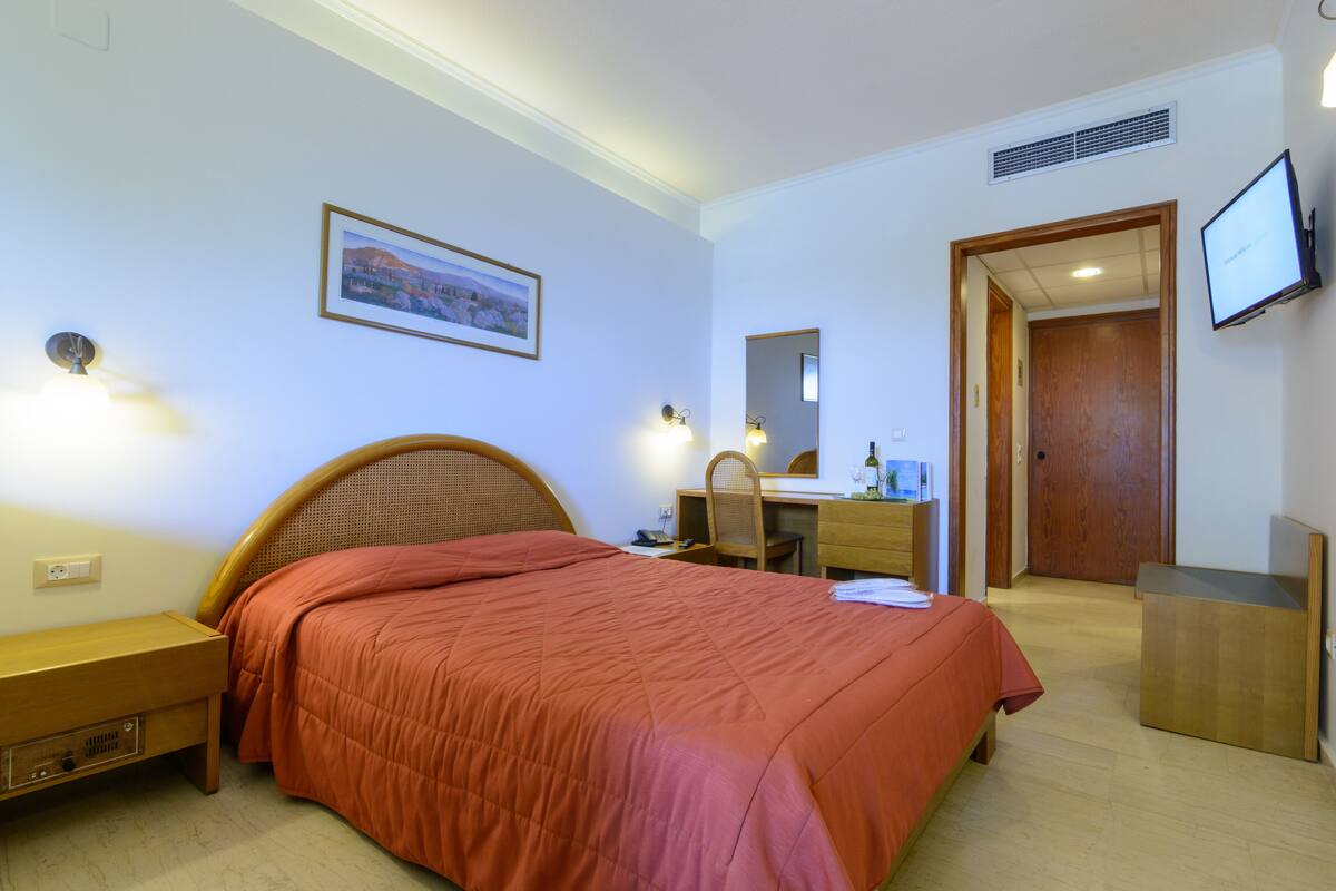 A hotel room with a bed, bedside table, desk, chair, mirror, TV, and a door.