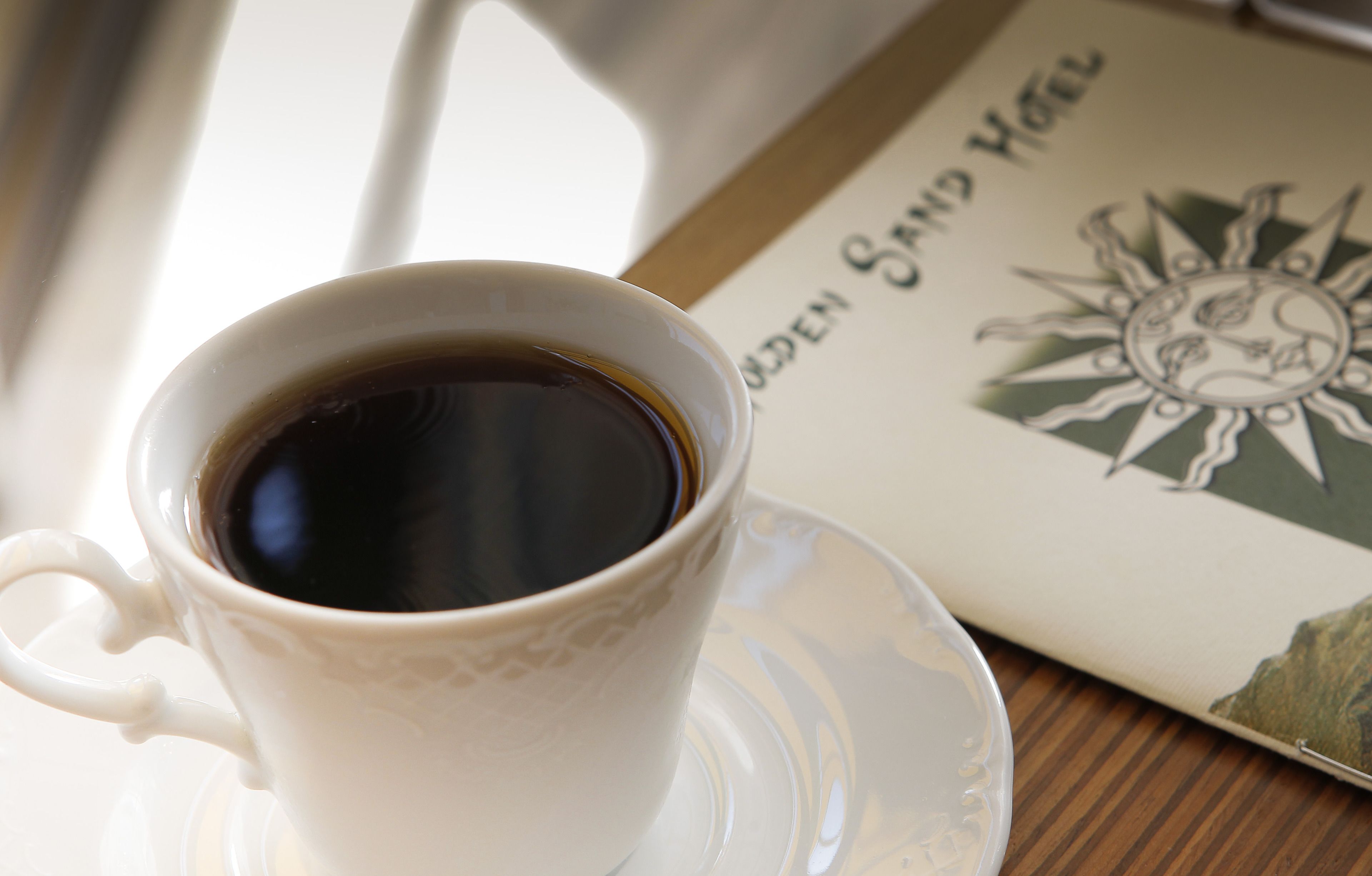 A cup of coffee on a saucer with a book titled "Golden Sand Hotel" beside it.
