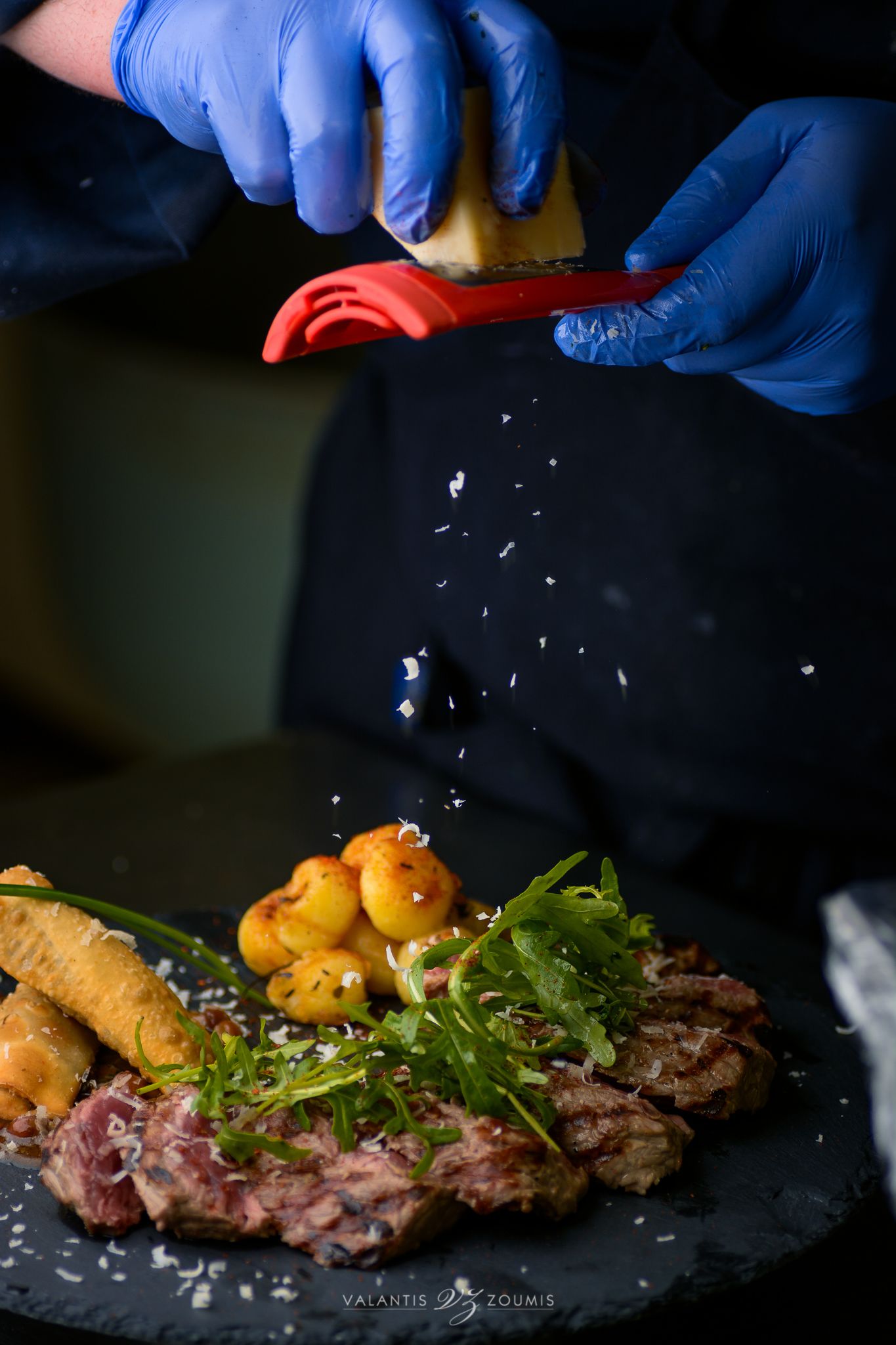 A person in blue gloves preparing food on a black plate, including meat, greens, and potatoes.