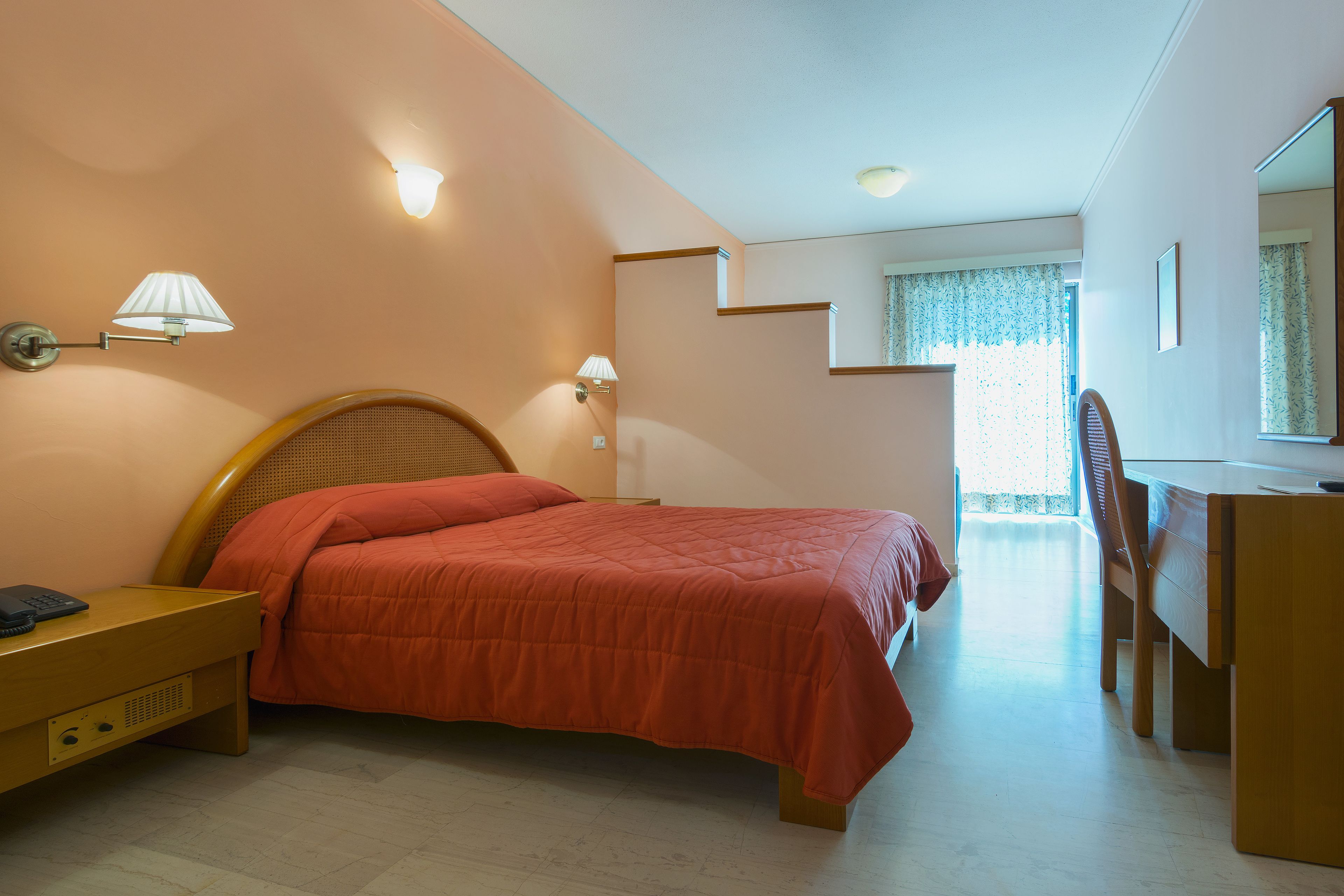 A bedroom with a bed, bedside table, lamp, desk, chair, and a view of a balcony.